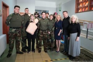 Grupa ludzi pozująca do zdjęcia w korytarzu budynku, z kilkoma osobami w mundurach moro na pierwszym planie. Większość osób ma jasną skórę. Po lewej stoją dwaj wysocy mężczyźni w mundurach w kamuflażu, w zielonych bluzach i czarnych butach. Obok nich stoi kobieta w czarnym stroju i okularach, trzymająca fantom do ćwiczeń pierwszej pomocy (tors z głową w jasnym, beżowym kolorze). Za nią widać osobę w hełmie i okularach ochronnych oraz kilka innych osób częściowo zasłoniętych. W centrum stoją dwie osoby w mundurach moro (zielone bluzy, pasy, czarne buty), jedna z ciemnymi włosami związanymi z tyłu, druga z krótkimi włosami i okularami. W tle jest też młoda osoba z kręconymi włosami oraz ktoś z różowym pasmem we włosach i okularami. Po prawej stronie stoją trzy osoby w cywilnych ubraniach: kobieta w granatowej sukience, mężczyzna w czarnej koszuli i jeansach oraz starsza kobieta z długimi siwymi włosami, w czarnym swetrze i szarej plisowanej spódnicy. W pomieszczeniu widać płytki na podłodze, drzwi po lewej, tablicę lub gablotę na ścianie po prawej oraz znaki i tablice informacyjne w tle. Na dole zdjęcia jest pasek ostrzegawczy w żółto-czarne pasy. W prawym dolnym rogu znajduje się podpis fotografa: „fot. Magdalena Seredyńska/Uniwersytet Szczeciński”.