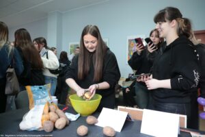 Grupa młodych kobiet, przy stole z ziemniakami i miską. W centrum stoi młoda kobieta z długimi prostymi brązowymi włosami, w czarnej bluzie. Trzyma ziemniaka i obiera go nad dużą zieloną miską. Na stole leży kilka nieobranych ziemniaków oraz siatka z ziemniakami z widocznym napisem „ZIEMNIAKI”. Obok są też drobne przedmioty kuchenne i białe kartki/karteczki na stojakach. Po prawej stoi inna młoda kobieta w czarnej bluzie, z ciemnymi włosami związanymi w kucyk, uśmiechnięta; w tle za nią dwie osoby trzymają telefony, jakby robiły zdjęcia lub nagrywały. Po lewej widać kilka osób stojących blisko siebie, część odwrócona plecami. Na ścianie z tyłu wiszą kolorowe obrazy. W lewym dolnym rogu jest podpis: „fot. Magdalena Seredyńska/Uniwersytet Szczeciński”.