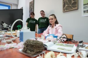 Trzy młode kobiety, przy dużym stole z rozłożonymi przedmiotami do zajęć. Na pierwszym planie jest stół z różnymi rzeczami: biała pompa lub urządzenie z wężem, małe miseczki, zielony spryskiwacz, przewody, otwarta książka z ilustracjami oraz dekoracje w postaci jasnoróżowych kwiatów. Po lewej stronie w tle stoi mikroskop i duży czarny monitor. Dwie kobiety stoją z tyłu i uśmiechają się; mają na sobie zielone koszulki z białymi napisami. Na koszulkach widać m.in. słowa „HYDROBIOLOGY” oraz „OCHRONA I INŻYNIERIA ŚRODOWISKA PRZYRODNICZEGO”. Trzecia kobieta siedzi bliżej aparatu przy stole; ma jasną cerę, ciemne włosy związane wysoko i różową marynarkę, patrzy w stronę aparatu. Na ścianie wiszą dekoracyjne panele z liściastym wzorem, a po prawej stronie jest plakat z małymi zdjęciami. W prawym dolnym rogu widoczny jest napis: „fot. Magdalena Seredyńska/Uniwersytet Szczeciński”.