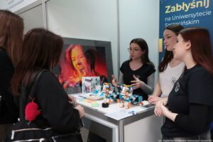 Stoisko informacyjne na wydarzeniu uczelnianym, przy którym stoi grupa młodych kobiet i rozmawia. Na blacie leżą małe modele robotów z klocków w kolorach biały, niebieski i pomarańczowy, a także ulotki i drobne materiały promocyjne. W tle widać duży ekran z kolorowym zdjęciem twarzy osoby krzyczącej lub śpiewającej w czerwono-pomarańczowym świetle. Po prawej stronie znajduje się pionowy baner z napisem „Zabłyśnij na Uniwersytecie Szczecińskim”. W prawym dolnym rogu jest podpis fotograficzny: „fot. Magdalena Seredyńska/Uniwersytet Szczeciński”.