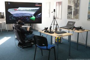 Stanowisko symulatora jazdy w pomieszczeniu, z dużym telewizorem na stojaku i osobą siedzącą w fotelu-kokpicie z kierownicą. Na ekranie telewizora wyświetlona jest gra wyścigowa z widokiem z wnętrza samochodu na torze; widać wirtualną kierownicę i zieloną rękawicę kierowcy oraz elementy interfejsu (m.in. minimapę i wskaźniki). Osoba ma na głowie gogle VR i siedzi w czarnym fotelu symulatora. Z przodu stoi niebieskie krzesło. Po prawej stronie są dwa stoły z laptopem, klawiaturą i różnym osprzętem; na blacie leżą także kontrolery/joysticki, przewody i butelka z rozpylaczem. Obok stoi statyw z małym urządzeniem na górze. Na ścianie wiszą duże obrazy/plakaty. W prawym dolnym rogu zdjęcia jest podpis: „fot. Magdalena Seredyńska/Uniwersytet Szczeciński”.