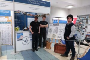 Stoisko promocyjne Uniwersytetu Szczecińskiego z trzema mężczyznami stojącymi obok ekspozycji. W tle są niebiesko-białe ścianki i banery z napisem „UNIWERSYTET SZCZECIŃSKI” oraz „Wydział Ekonomii, Finansów i Zarządzania”. Na ladzie stoi laptop i miska z zielonymi jabłkami. Po lewej stronie widać dużą tablicę z wieloma małymi karteczkami oraz rząd cyfr od 4 do 10. Po prawej stronie stoi sztaluga z planszą zatytułowaną „Gospodarka Przestrzenna” z rysunkami architektonicznymi. Obok jest tablica/ścianka z wieloma małymi obrazkami i magnesami. Na podłodze leżą brązowe torby podróżne. Osoby: po lewej mężczyzna w czarnej koszulce i czapce z daszkiem, z okularami i krótkim zarostem; w środku mężczyzna w czarnej koszulce, z rękami założonymi; po prawej mężczyzna w czarno-szarej kurtce typu varsity i czarnym meloniku. Widoczny podpis w lewym dolnym rogu: „fot. Magdalena Seredyńska/Uniwersytet Szczeciński”.