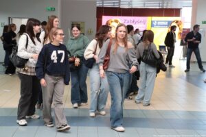 Grupa młodych osób na korytarzu uczelni. Większość to młode kobiety o jasnej karnacji, ubrane na co dzień w dżinsy i bluzy, część niesie torby na ramię i plecaki. Na pierwszym planie po lewej idą dwie dziewczyny: jedna w granatowej bluzie z dużym białym numerem „47” i okularach, druga w jasnej bluzie z nadrukiem. Na środku przodu idzie dziewczyna w szarej bluzie odsłaniającej jedno ramię i szerokich dżinsach. W tle widać więcej studentów oraz stoiska promocyjne z banerami; na jednym banerze jest czytelny napis „Zabłyśnij”, a na innym „Uniwersytet Szczeciński” i adres „kandydaci.usz.edu.pl”. W prawym dolnym rogu zdjęcia jest podpis: „fot. Magdalena Seredyńska/Uniwersytet Szczeciński”.