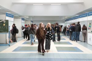 Jasny korytarz w budynku uczelni z rzędami stoisk informacyjnych po obu stronach i grupami ludzi między nimi. W centrum kadru dwie młode kobiety idą w stronę aparatu; jedna ma bardzo długie jasnobrązowe włosy i brązową, wzorzystą kurtkę, druga ma jasne włosy, okulary i czarną bluzę z białym wzorem. Po bokach stoją studenci, część rozmawia przy stoiskach, część trzyma kartki lub torby. Nad stoiskami wiszą niebiesko-białe tablice z nazwami wydziałów, m.in. „Wydział Humanistyczny” oraz „Wydział Nauk Społecznych”. Podłoga jest w duże kwadraty w odcieniach bieli, szarości i niebieskiego, a sufit ma długie świetlówki. W prawym dolnym rogu widoczny jest podpis: „fot. Magdalena Seredyńska / Uniwersytet Szczeciński”.