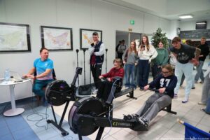 Pomieszczenie wewnątrz budynku z dwoma ergometrami wioślarskimi ustawionymi obok siebie na podłodze z jasnych płytek. Na pierwszym planie dwie osoby siedzą na maszynach i „wiosłują”, trzymając uchwyty z linką. Osoba po prawej ma jasne włosy, okulary i ciemną bluzę z dużym numerem „47”. Osoba bardziej w środku ma brązowe włosy i bordową bluzę. Po lewej stronie mężczyzna w niebieskiej koszulce siedzi przy małym okrągłym stoliku z otwartym laptopem; na stole stoi też butelka wody i drobne przedmioty. Z maszyn do wiosłowania wychodzą przewody prowadzące w stronę stołu. W tle stoi grupa młodych osób obserwujących wydarzenie; część trzyma telefony. Na ścianie wiszą trzy duże mapy w czarnych ramach. Widać też stojaki (prawdopodobnie do sprzętu nagraniowego) i zielony znak wyjścia ewakuacyjnego nad drzwiami. W prawym dolnym rogu jest mały napis ze stopką fotograficzną, ale jest zbyt drobny, by pewnie przepisać.