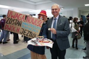 Wnętrze budynku z grupą ludzi, na pierwszym planie starszy mężczyzna w garniturze i postać w stroju dużego misia. Mężczyzna ma siwe włosy, uśmiecha się i trzyma w dłoni okrągłe ciasteczko. Miś ma brązowe futro, czerwoną czapkę z żółtym paskiem i granatowy płaszcz; przed nim jest taca z różnymi słodyczami, w tym podłużnymi ciasteczkami, kolorowymi żelkami oraz pudełkiem z motywem brytyjskiej flagi. Obok misia trzymany jest duży kartonowy transparent z napisem: „UWAGA! NOWOŚĆ ZOSTAŃ NAUCZYCIELEM TŁUMACZEM DWÓCH JĘZYKÓW”. W tle widać osoby stojące i rozmawiające, a także kamerę lub sprzęt nagraniowy po prawej stronie. W prawym dolnym rogu jest podpis: „fot. Magdalena Seredyńska/Uniwersytet Szczeciński”.