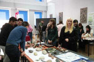 Grupa młodych osób w jasnej sali przy długim stole z okazami kamieni i minerałów. Na blacie leżą różne skały o różnych kształtach i kolorach, kartki z opisami, butelka z wodą, kilka pojemników i mała gablotka z próbkami; z przodu po prawej widać duży arkusz z rysunkami lub mapami. Kilka osób stoi i pochyla się nad eksponatami, jak na zajęciach lub prezentacji. W tle są wysokie okna z niebieskimi zasłonami, tablica lub ekran oraz ścienne dekoracje; po prawej kolejny stół z białymi obiektami przypominającymi kości lub czaszki zwierząt i osoby pracujące przy nim. Na ścianie po prawej jest plakat z drobnym tekstem, ale treść nie jest czytelna.