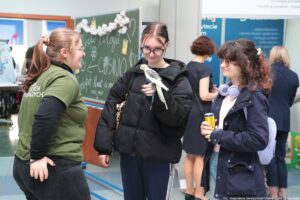 Trzy młode kobiety rozmawiające w korytarz. Po lewej stronie stoi kobieta w zielonej koszulce i czarnych spodniach, z jasnobrązowymi włosami związanymi w kucyk; jest zwrócona bokiem do pozostałych. W centrum stoi kobieta w czarnej, puchowej kurtce i okularach, z ciemnymi włosami; na dłoni trzyma małą papugę nimfę (szaro-białe pióra, żółta głowa z pomarańczową plamką na policzku). Po prawej stoi kobieta w granatowej kurtce, z kręconymi ciemnymi włosami i okularami; na szyi ma duże fioletowe słuchawki i w dłoni trzyma żółtą puszkę napoju, a na plecach jasny plecak. W tle widać tablicę z napisami kredą, ozdobioną białymi kwiatami, oraz kilka osób stojących przy niebieskich planszach informacyjnych. W prawym dolnym rogu jest podpis: „fot. Magdalena Seredyńska/Uniwersytet Szczeciński”.