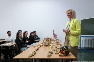 Sala wykładowa z grupą studentów oraz prowadzącą stojącą przy stole z kośćmi. Prowadząca to dorosła kobieta o jasnej skórze i jasnych, kręconych włosach, ubrana w jaskrawożółtą marynarkę i jasne spodnie; uśmiecha się i trzyma w dłoniach mały przedmiot. Po lewej stronie siedzi kilka osób w ławkach, skierowanych w stronę prowadzącej, jak podczas zajęć. Na długim stole na pierwszym planie leżą rozłożone elementy szkieletu (kręgosłup z żebrami, kości kończyn, fragmenty miednicy) oraz czaszka; obok znajduje się też kolorowy model czaszki i anatomiczny model mięśni. W tle po prawej widać tablicę i grzejnik. W prawym dolnym rogu jest podpis: „fot. Magdalena Seredyńska/Uniwersytet Szczeciński”.
