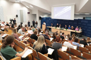 Duża sala wykładowa wypełniona osobami piszącymi dyktando. W rzędach drewnianych, amfiteatralnie ustawionych ławek siedzą uczestnicy z kartkami i długopisami, pochylają się nad notatkami. Na dole, przy przedniej ścianie, stoi długi stół prezydialny, za którym siedzi kilkanaście osób w garniturach i eleganckich strojach, przed nimi stoją zielone butelki z wodą i bukiet kwiatów na środku stołu. Z lewej strony przy mównicy stoi kobieta czytająca tekst do mikrofonu. Za stołem i mównicą znajduje się granatowa ścianka z powtarzającym się białym logo i napisem „UNIWERSYTET SZCZECIŃSKI”. Nad nią wisi duży ekran z jasnoniebieskim slajdem, na którym widoczne są słowa „DYKTANDO UNIWERSYTECKIE” oraz godzina „11:00”; po prawej na ekranie znajduje się odręczny podpis. Po lewej stronie sali ustawione są kamery na statywach, obok których stoją osoby obserwujące wydarzenie. Ściany są jasne, na nich wiszą prostokątne panele akustyczne o geometrycznym wzorze. Całość sprawia wrażenie oficjalnego wydarzenia akademickiego na Uniwersytecie Szczecińskim.