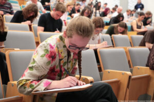 Studentka w kolorowym, kwiecistym żakiecie siedząca w sali wykładowej i pochylona nad kartką. Ma długie, jasnobrązowe włosy zaplecione w warkocz i okulary z ciemnymi oprawkami. Pisze długopisem na białej kartce leżącej na małym, rozkładanym blaciku przymocowanym do fotela. Wokół niej widać rzędy szaro‑drewnianych krzeseł wykładowych. W tle wiele innych osób, młodych i starszych, również skupionych na pisaniu, każda z kartką i długopisem. Atmosfera przypomina egzamin lub konkurs odbywający się w dużej auli. W prawym dolnym rogu znajduje się drobny napis z informacją o autorce zdjęcia i uczelni.