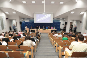 Sala wykładowa z amfiteatralnie ustawionymi rzędami ławek wypełnionych osobami piszącymi. Na dole, przed słuchaczami, stoi długi stół prezydialny, za którym siedzi osiem osób w oficjalnych strojach; na stole stoją mikrofony, kilka zielonych butelek z wodą i bukiet kwiatów pośrodku. Po lewej stronie stołu znajduje się mównica, przy której stoi osoba czytająca z kartki; obok ustawione są statywy z kamerami skierowanymi na przód sali. Tło sceny tworzą ciemnoniebieskie ścianki z białymi napisami „Uniwersytet Szczeciński” i herbami uczelni. Nad głowami prelegentów wisi biały ekran projekcyjny z wyświetlonym slajdem w odcieniach fioletu; widać duży napis „DYKTANDO” oraz logotyp Uniwersytetu Szczecińskiego i datę „22.04.2024, 11:00”. W górnej części kadru widać jasny sufit z szeregiem prostokątnych lamp. Po bokach sali, przy ścianach, stoją pojedyncze osoby obserwujące wydarzenie oraz sprzęt nagłośnieniowy. Uczestnicy w ławkach pochylają się nad kartkami, pisząc, większość ma na sobie codzienne, raczej jasne lub stonowane ubrania.