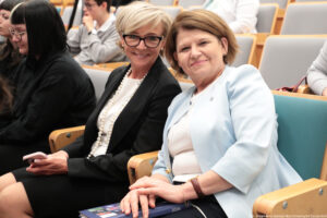 Dwie kobiety siedzące obok siebie w auli wykładowej. Obie są w średnim wieku, mają jasną karnację i krótkie, zadbane fryzury. Kobieta po lewej ma krótko obcięte, jasny blond włosy zaczesane na bok, nosi ciemne okulary w czarnych oprawkach, biały top lub bluzkę, długi naszyjnik z dużych pereł i czarną marynarkę oraz ciemną spódnicę. W dłoniach trzyma smartfon o jasnej obudowie, na kolanach ma założone nogi. Uśmiecha się w stronę aparatu. Kobieta po prawej ma jasnobrązowe włosy do linii podbródka, ułożone w delikatny bob. Ma na sobie jasny, prawdopodobnie biały top, na to jasnoniebieską marynarkę i ciemniejsze spodnie lub spódnicę. Na lewym nadgarstku ma brązowy pasek zegarka i cienką bransoletkę, na palcu obrączkę. Dłonie opiera na książce leżącej na kolanach; na grzbiecie książki widać rok „2026” i kolorową okładkę, ale tytuł nie jest czytelny. Kobieta patrzy w stronę aparatu i uśmiecha się łagodnie. Obie siedzą w wygodnych fotelach z drewnianymi podłokietnikami i tapicerką w odcieniach jasnoszarym i turkusowym, co sugeruje salę konferencyjną lub uniwersytecką. W tle widać kilka innych osób siedzących w rzędach, ale są mniej wyraźne. Na samym dole po prawej stronie jest drobny, biały napis z podpisem zdjęcia, jednak ze względu na mały rozmiar nie da się go przeczytać w całości.