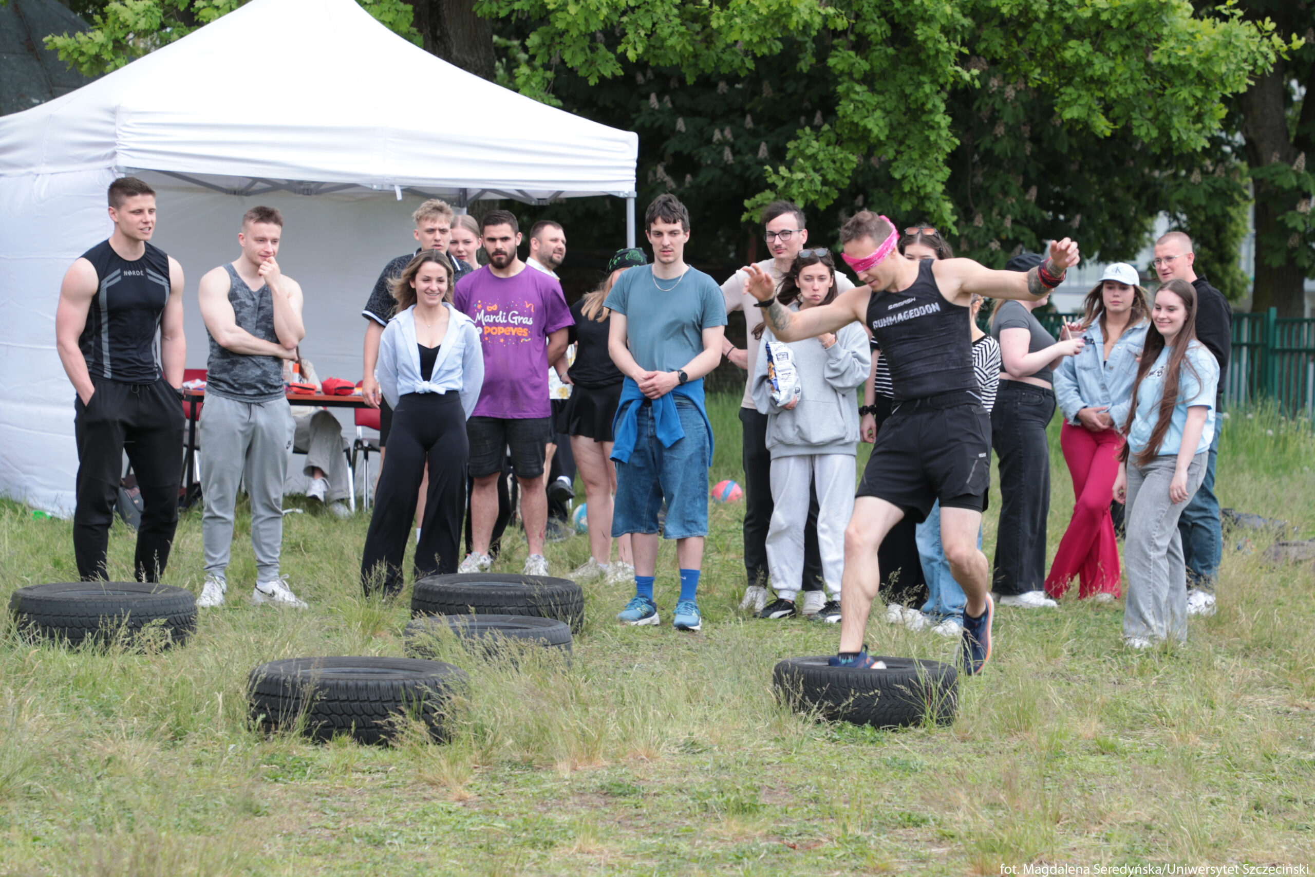 Grupa młodych ludzi na świeżym powietrzu podczas zawodów sportowych. Na pierwszym planie po prawej stronie biegnie mężczyzna o jasnej skórze w czarnej koszulce bez rękawów i czarnych spodenkach, z różową opaską na oczach i nadgarstkach. Stawia stopę na leżącej na trawie oponie, a ręce ma uniesione dla utrzymania równowagi. Przed nim i za nim w trawie leży kilka dużych, ciemnych opon ułożonych w rzędzie jako przeszkody terenowe. W tle stoi kilkanaście osób, większość w swobodnych sportowych ubraniach: dresach, T‑shirtach, legginsach. Część się uśmiecha, część obserwuje w skupieniu. Po lewej stronie widoczny jest biały namiot plenerowy z ławką i stołem w środku. Za ludźmi rosną zielone drzewa, co sugeruje teren parku lub boiska trawiastego. W dolnym prawym rogu znajduje się mały biały napis z informacją o autorce zdjęcia i uczelni.
