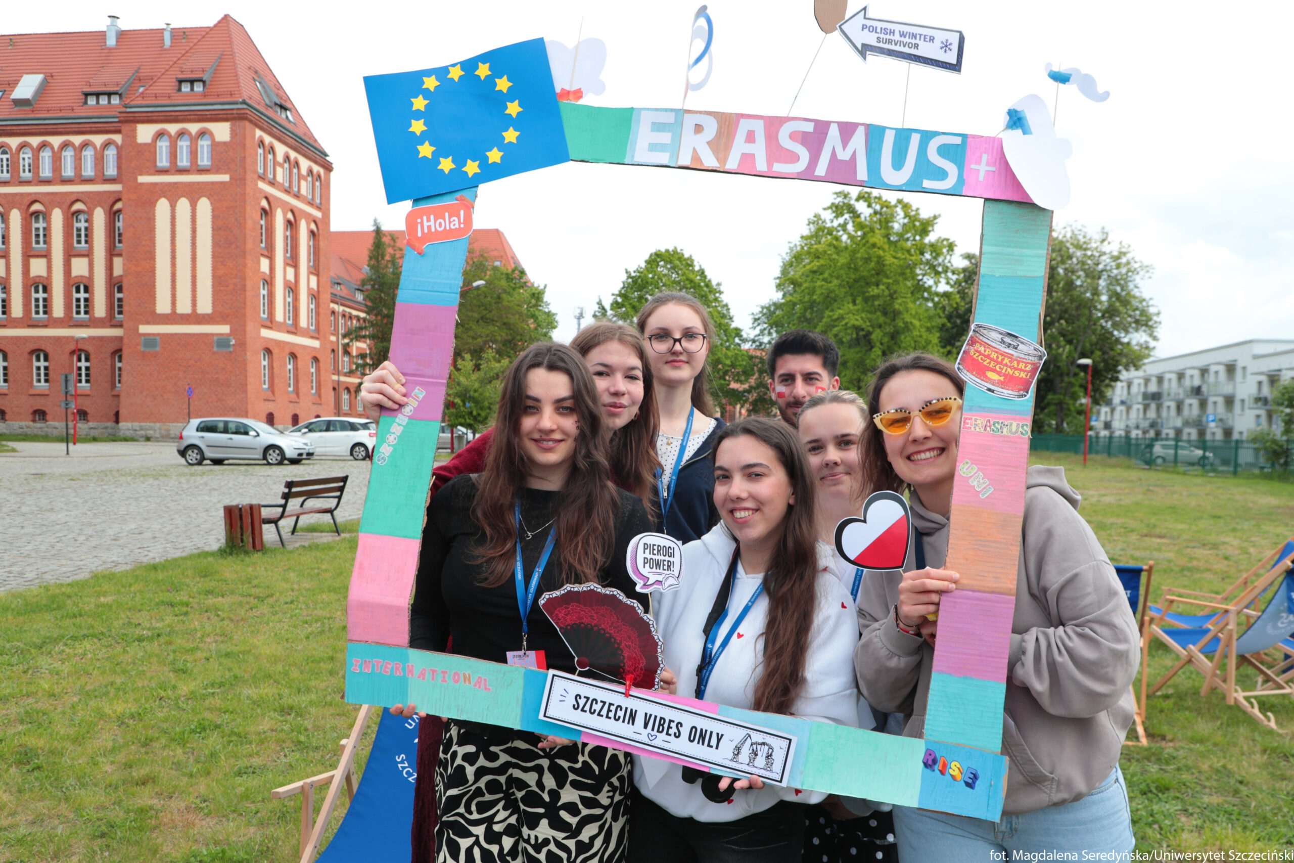 Grupa młodych osób stojąca razem w kolorowej ramce z napisem „ERASMUS+”. Na pierwszym planie widać siedem osób, wszystkie uśmiechnięte, w większości o jasnej lub średniej karnacji, w wieku licealno‑studenckim. Kilka z nich ma na szyi niebieskie smycze z identyfikatorami. Trzymana jest duża prostokątna ramka z tektury, pomalowana w pasy turkusu, różu, pomarańczu i fioletu. Na górze ramki widnieje napis „ERASMUS+” w dużych, kolorowych literach; przyczepiona jest też niebieska flaga Unii Europejskiej z żółtymi gwiazdami oraz pomarańczowa chmurka z napisem „¡Hola!”. Na dole ramki znajduje się biały pasek z czarnym napisem „SZCZECIN VIBES ONLY” i rysunkami, a na lewym pionowym boku mały napis „INTERNATIONAL”. Na prawym boku są niewielkie ozdoby, w tym naklejka z napisem „ERASMUS” oraz obrazek słoika „Paprykarz Szczeciński”. Kilka osób trzyma dodatkowe rekwizyty: jedna osoba wachlarz i tabliczkę z napisem „PIEROGI POWER!”, inna kartonowe serce w barwach flagi Polski. Jedna z dziewczyn ma żółte, trójkątne okulary przeciwsłoneczne. W tle po lewej stronie znajduje się duży czerwony, ceglany budynek o ozdobnej fasadzie, na trawniku stoją ławki i zaparkowane samochody. Po prawej stronie widać więcej zieleni oraz nowsze, jasne budynki mieszkalne. Całość sprawia wrażenie plenerowego wydarzenia przy uczelni.