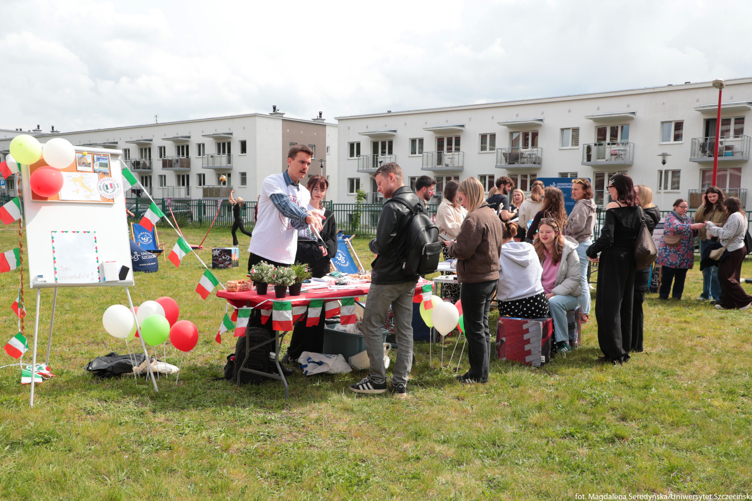 Plenerowy piknik na trawniku przed blokami mieszkalnymi. Na pierwszym planie stoi stół przykryty czerwonym obrusem, udekorowany małymi flagami w barwach zielono‑biało‑czerwonych oraz kilkoma balonami w tych samych kolorach. Na stole leżą ulotki, talerze z jedzeniem i trzy małe doniczki z roślinami. Za stołem stoi młody mężczyzna o jasnej karnacji w białej koszulce z nadrukiem, który gestykuluje w kierunku rozmówcy, obok niego stoi młoda kobieta. Przed stołem stoi mężczyzna z plecakiem i kilka innych osób, które rozmawiają i oglądają przygotowane rzeczy. Po lewej stronie ustawiona jest biała tablica na sztaludze, ozdobiona balonami i małymi flagami, z kilkoma karteczkami i plakatami przyczepionymi u góry. Pod tablicą leżą torby i inne drobiazgi. W głębi widać większą grupę ludzi zgromadzonych przy kolejnych stołach oraz niskie, białe budynki mieszkalne z balkonami. Nad całą sceną jest pochmurne, ale dość jasne niebo, sugerujące dzienną porę.