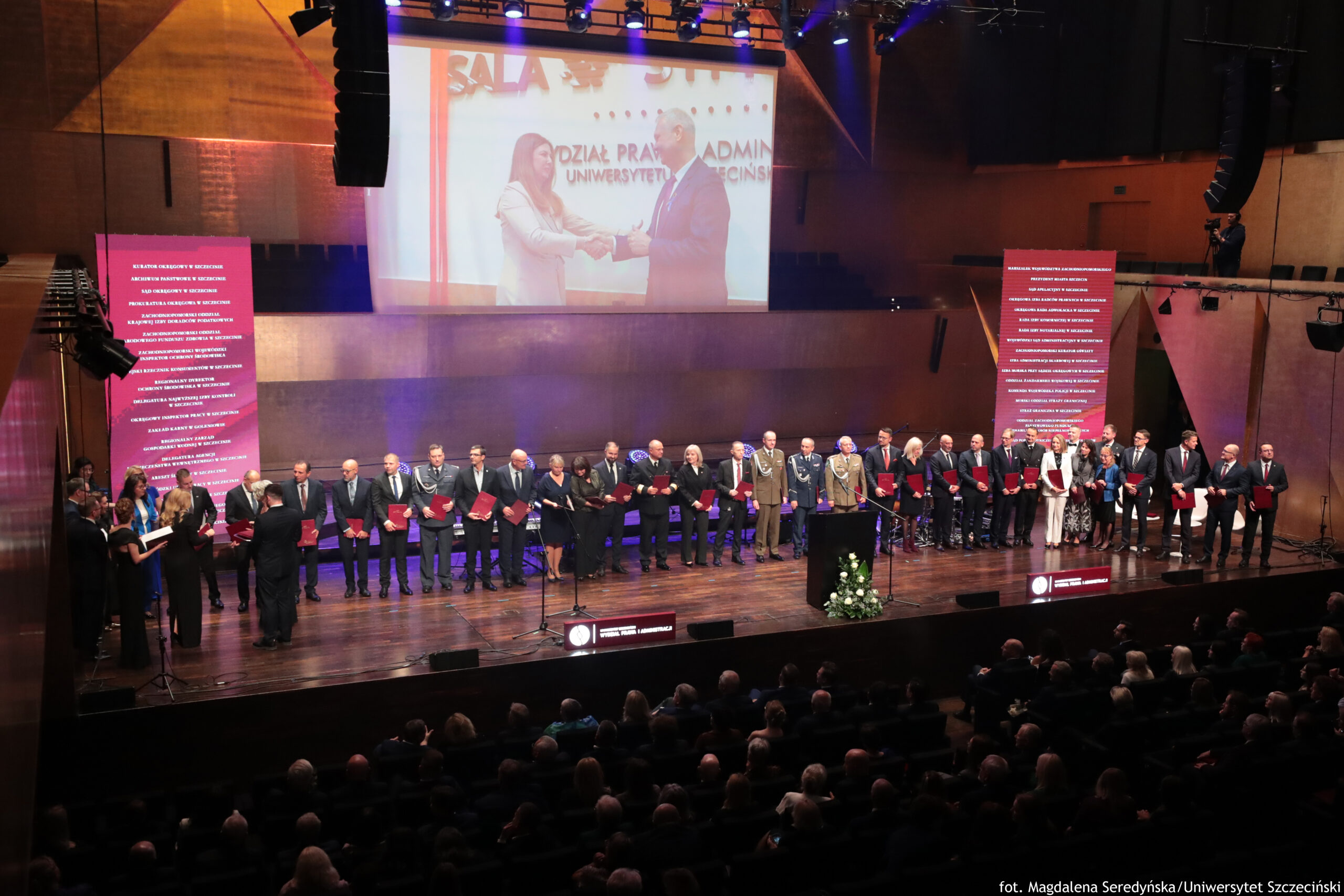 Gala 40-lecia Wydziału Prawa i Administracji Uniwersytetu Szczecińskiego. Na scenie w jednym rzędzie stoi 30-osobowa grupa ludzi powołanych do Rady Interesariuszy Wydziału Prawa i Administracji. Po prawej i lewej dwa pionowe ekrany z nazwiskami. Pośrodku poziomy ekran z filmem.