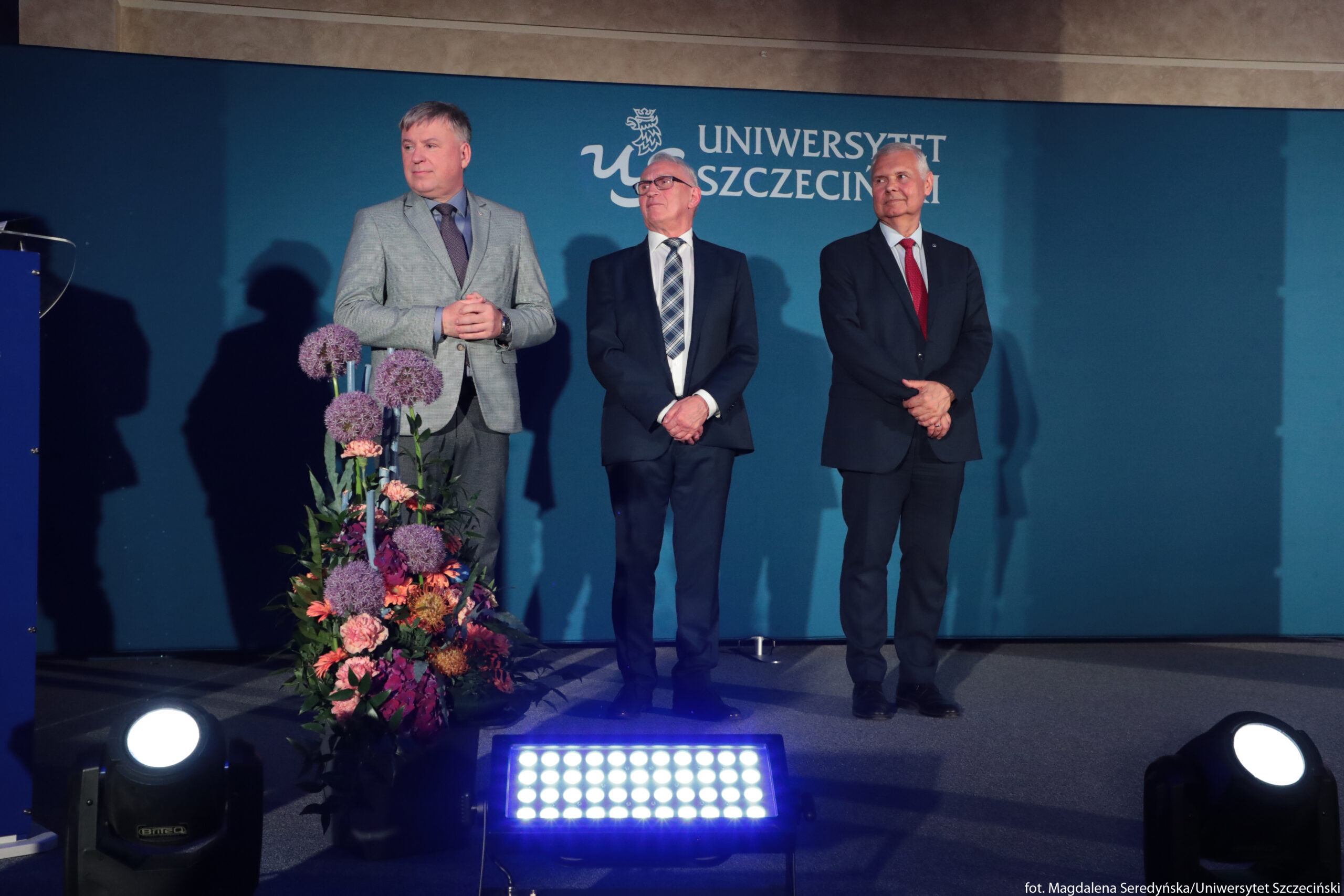 Trzech mężczyzn w garniturach stojących na tle ściany z napisem „Uniwersytet Szczeciński”. Po lewej mężczyzna w jasnoszarym garniturze, z fioletowym krawatem, krótko ostrzyżony, o jasnych włosach; ma złożone dłonie na wysokości brzucha i patrzy lekko w lewo. W środku stoi starszy mężczyzna w ciemnym garniturze, białej koszuli i krawacie w granatowo‑szarą kratę; ma jasne włosy zaczesane do tyłu, okulary i patrzy lekko w górę. Po prawej stoi mężczyzna w ciemnym garniturze, białej koszuli i czerwonym krawacie; ma siwe włosy, ręce splecione przed sobą i jest lekko uśmiechnięty. Za nimi jednolite, ciemnoturkusowe tło z logotypem i napisem „Uniwersytet Szczeciński” w białym kolorze. Przed mężczyzną po lewej znajduje się duży, wysoki bukiet w wazonie z różnokolorowymi kwiatami: kulistymi fioletowymi kwiatostanami, różowymi goździkami i innymi kwiatami w odcieniach pomarańczu, różu i fioletu, z zielonymi liśćmi. Na podłodze z przodu ustawione są jasne reflektory sceniczne, w tym prostokątna listwa z wieloma niebieskawymi diodami LED, skierowana w górę. Oświetlenie jest sceniczne, z wyraźnymi cieniami sylwetek mężczyzn na tle ściany.