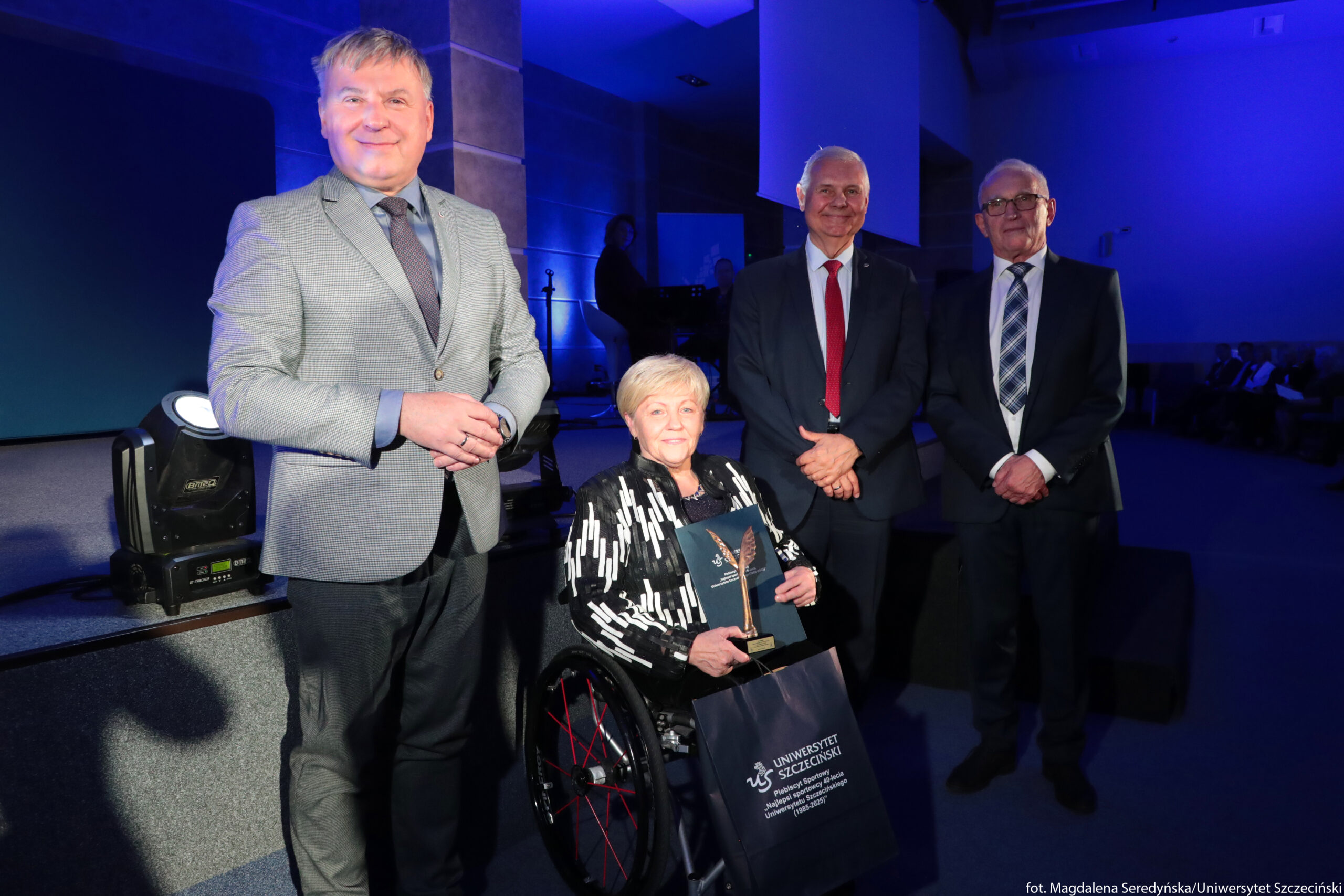 Cztery dorosłe osoby w oficjalnych strojach podczas uroczystości w dobrze oświetlonej sali. Po lewej stoi mężczyzna w jasnoszarej marynarce, błękitnej koszuli i ciemnym krawacie, z rękami złożonymi przed sobą, uśmiechnięty. Obok, na pierwszym planie, znajduje się starsza kobieta o jasnych, krótkich włosach, siedząca na wózku. Ubrana jest w czarno‑białą, wzorzystą marynarkę. Na kolanach trzyma statuetkę w kształcie skrzydła lub skrzydeł na prostokątnej podstawie oraz granatową teczkę lub okładkę. Przed wózkiem wisi granatowa torba z białym nadrukiem „Uniwersytet Szczeciński” i dalszym tekstem poniżej. Po prawej stronie kobiety stoją dwaj starsi mężczyźni w ciemnych garniturach. Mężczyzna bliżej środka ma czerwoną krawatę i biały krawatnik koszuli lekko wystający ponad marynarkę, również się uśmiecha. Mężczyzna najbardziej po prawej ma okulary, białą koszulę i krawat w niebiesko‑białą kratę, trzyma dłonie splecione przed sobą. Tło jest niebiesko podświetlone, scena wygląda jak akademia lub gala, w głębi widać zarys sprzętu scenicznego i sylwetki osób siedzących. W dolnym prawym rogu widoczny jest mały biały napis z przypisaniem zdjęcia: „fot. Magdalena Seredyńska/Uniwersytet Szczeciński”.