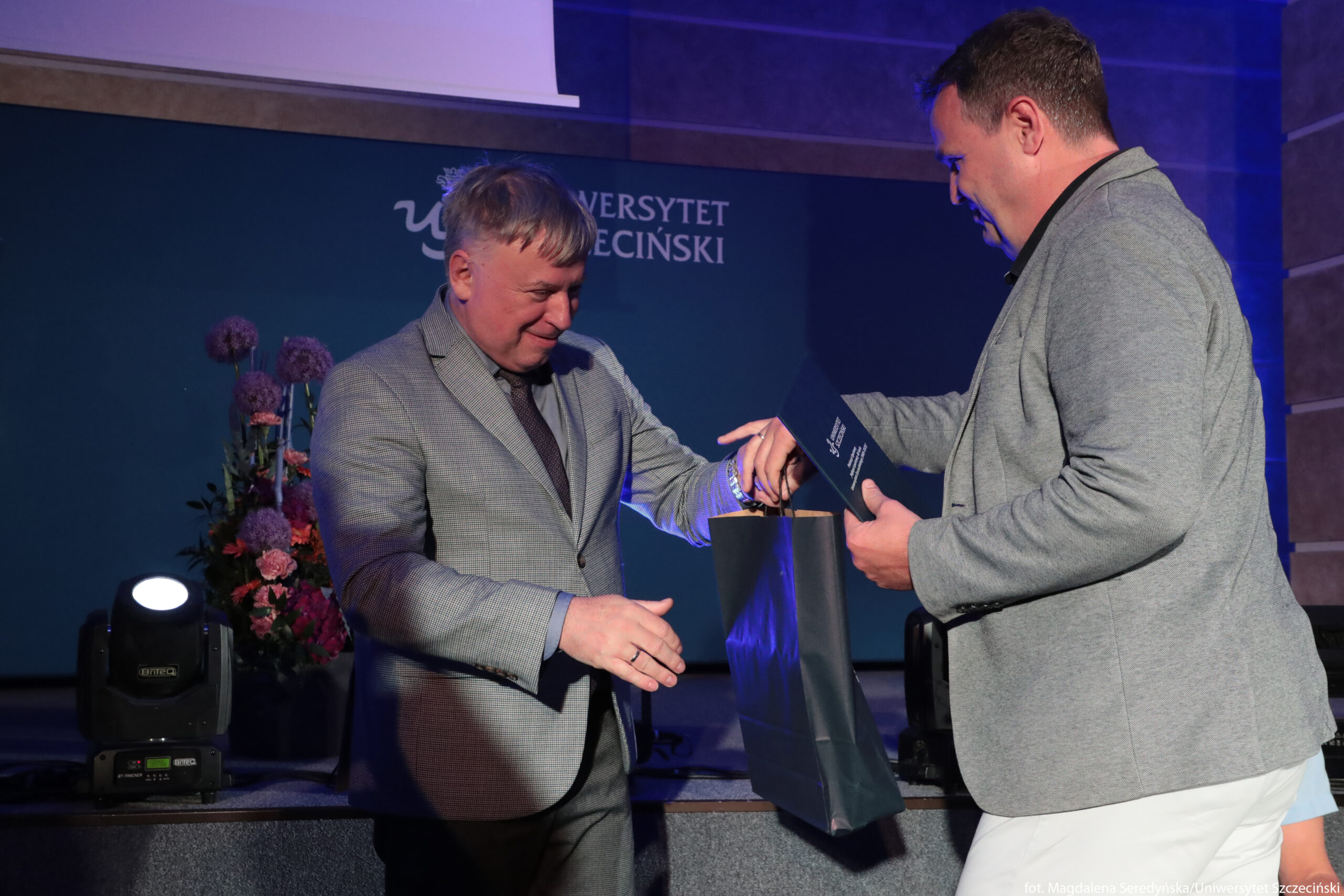 Dwaj mężczyźni w średnim wieku podczas wręczania nagrody na scenie. Po lewej stronie stoi mężczyzna o jasnej cerze, z krótkimi, siwymi włosami, ubrany w szarą marynarkę w drobną kratkę, koszulę i krawat w zbliżonym kolorze. Uśmiecha się i wyciąga lewą rękę, jakby miał przejąć prezent. Po prawej stronie stoi drugi mężczyzna o jasnej cerze, z krótkimi, ciemniejszymi włosami, ubrany w jasnoszarą marynarkę i jasne spodnie. W jednej ręce trzyma granatową teczkę lub dyplom, drugą podaje ciemną, elegancką torebkę prezentową z uchwytami ze sznurka, skierowaną w stronę mężczyzny po lewej. W tle widoczna jest ściana w odcieniach ciemnego turkusu, na której znajduje się biały napis „UNIWERSYTET SZCZECIŃSKI” oraz element graficzny z herbem uczelni. Z lewej strony za mężczyzną ustawione są wysokie kompozycje kwiatowe w odcieniach fioletu i różu. U dołu, po obu stronach sceny, stoją czarne reflektory sceniczne świecące jasnym światłem. Oświetlenie ma chłodny, niebieskawy ton, co podkreśla uroczysty, oficjalny charakter wydarzenia.