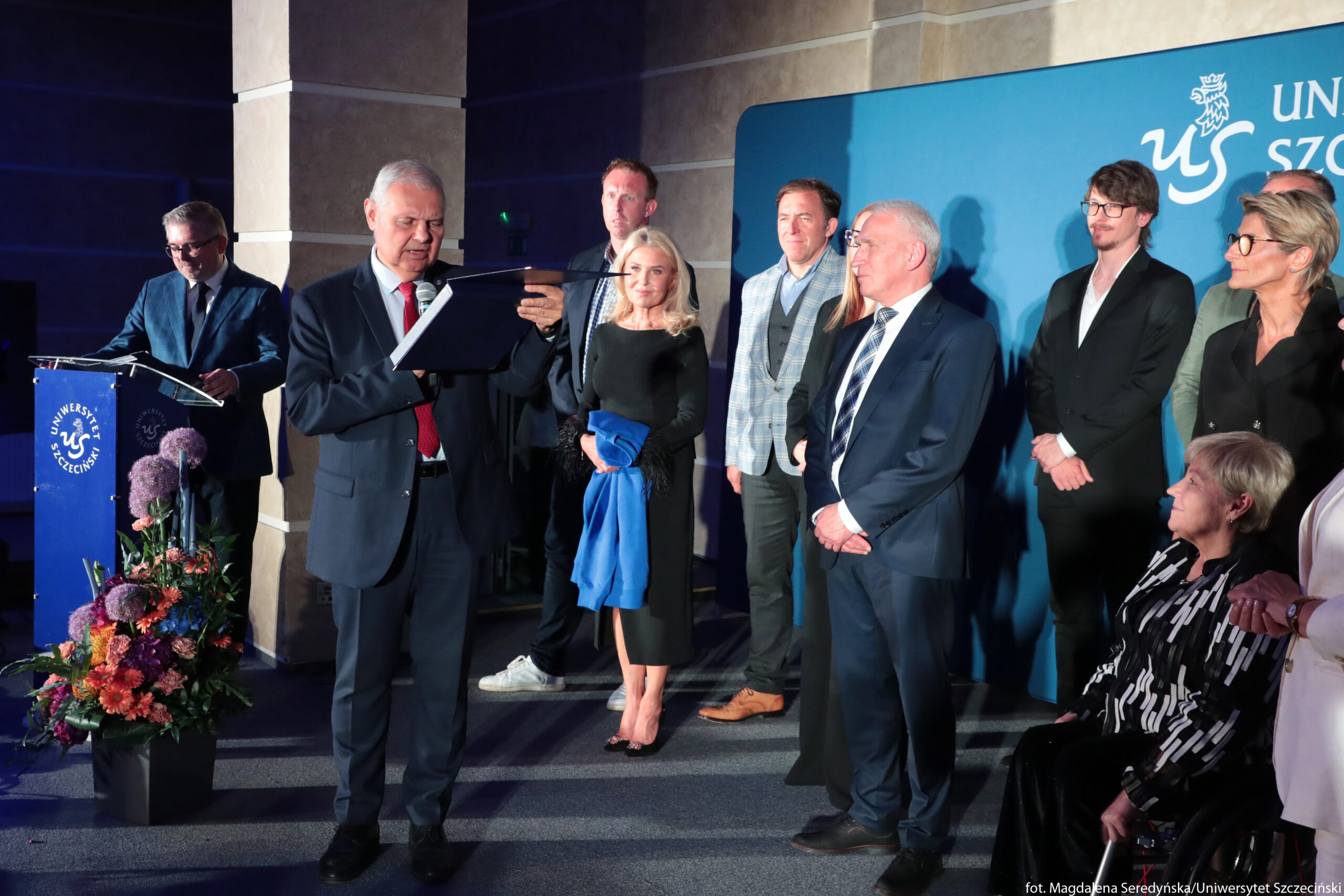 Grupa elegancko ubranych osób podczas oficjalnej uroczystości na Uniwersytecie Szczecińskim. Na pierwszym planie po lewej stronie stoi starszy mężczyzna o jasnych włosach w ciemnym garniturze z czerwonym krawatem; trzyma mikrofon i czyta z ciemnej teczki lub dyplomu. Za nim, przy mównicy z niebieskim frontem i białym logo uczelni, stoi inny mężczyzna w okularach, w granatowej marynarce; również ma przed sobą dokumenty. Pośrodku kadru stoi kilka osób w szeregu: kobieta w czarnej dopasowanej sukience do połowy łydki, z jasnymi rozpuszczonymi włosami, trzyma w rękach niebieski płaszcz; za nią stoi wysoki mężczyzna w ciemnym garniturze. Obok znajdują się kolejni mężczyźni w garniturach, w tym jeden w jasnoszarą kratę z ciemną kamizelką i brązowymi butami, inny starszy z siwymi włosami w granatowym garniturze i pasiastym krawacie. Dalej w prawo widać młodszego mężczyznę w czarnym garniturze, z ciemnymi włosami i okularami, oraz kobietę o krótkich, jasnych włosach w okularach, ubrana na ciemno. Na samym dole po prawej stronie siedzi starsza kobieta w czarno‑białej wzorzystej marynarce, na wózku inwalidzkim; obok niej częściowo widoczna jest osoba w jasnej marynarce. W tle po prawej stronie biegnie duża niebieska ściana z logo i napisem „Uniwersytet Szczeciński”, po lewej przy mównicy stoi wysoki stojak z kolorową kompozycją kwiatową w odcieniach różu, fioletu, czerwieni i pomarańczu.