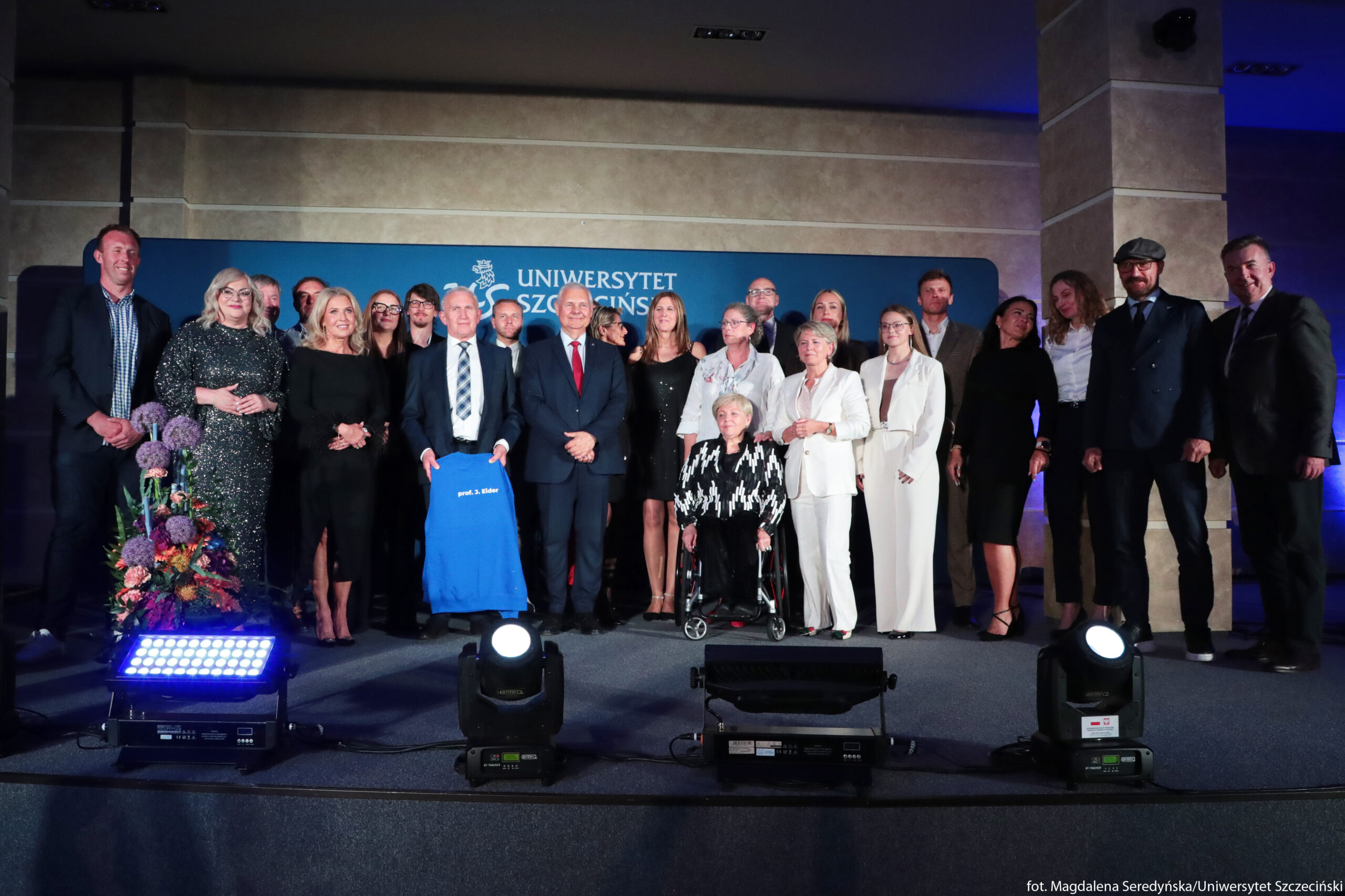 Grupa około dwudziestu pięciu osób stojąca na scenie na tle niebieskiej ścianki z napisem „Uniwersytet Szczeciński”. W centrum stoją starsi mężczyźni w garniturach; jeden z nich trzyma przed sobą niebieską bluzę z białym napisem „prof. J. Eider”. Przed nimi, lekko z prawej strony, siedzi starsza kobieta o siwych włosach na wózku, ubrana w czarno-białą wzorzystą marynarkę i czarne spodnie. Po obu stronach widać osoby w różnym wieku, ubrane odświętnie: kobiety głównie w sukienkach lub jasnych garniturach, mężczyźni w garniturach, marynarkach lub koszulach; po prawej jeden mężczyzna ma kaszkiet i brodę. Na samym przodzie stoją na podłodze duże reflektory sceniczne skierowane w stronę grupy; po lewej stronie przed grupą znajduje się wysoki, kolorowy bukiet kwiatów w fioletach, czerwieniach, różu i zieleni. Tło jest ciemne, oświetlone niebieskim światłem, z beżowymi filarami po lewej i prawej stronie sceny.