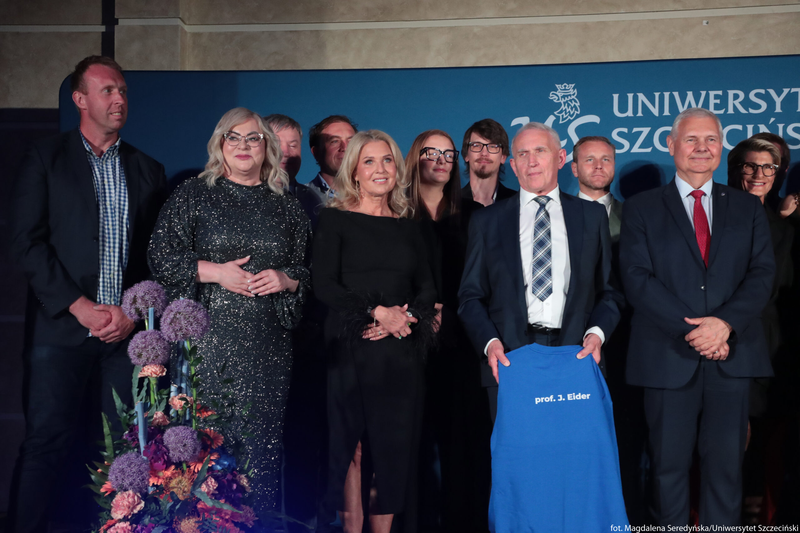 Grupa dorosłych osób stojących razem na tle niebieskiej ścianki z napisem „Uniwersytet Szczeciński”. W pierwszym rzędzie stoi pięć osób: po lewej mężczyzna w granatowej marynarce i koszuli w kratę, obok kobieta w ciemnej, błyszczącej sukni do ziemi z jasnymi, rozpuszczonymi włosami i okularami, dalej kobieta w czarnej dopasowanej sukni z dłuższym rękawem wykończonym piórami i rozpuszczonymi blond włosami. Obok niej stoi starszy mężczyzna w granatowym garniturze, białej koszuli i krawacie w niebiesko‑szarą kratę; trzyma przed sobą jasnoniebieską koszulkę z białym nadrukiem „prof. J. Eider”. Po prawej stronie stoi kolejny starszy mężczyzna w ciemnym garniturze, białej koszuli i czerwonym krawacie, z rękami skrzyżowanymi z przodu, uśmiechnięty. Za nimi widocznych jest kilka kolejnych osób w drugim rzędzie, częściowo przysłoniętych, w strojach wizytowych; wśród nich m.in. mężczyzna z krótką brodą i okularami, kobieta w dużych, ciemnych okularach oraz inni w marynarkach i koszulach. Na dole po lewej stronie zdjęcia znajduje się wysoka, kolorowa kompozycja kwiatowa z fioletowymi kulistymi kwiatami i pomarańczowo‑różowymi dodatkami.