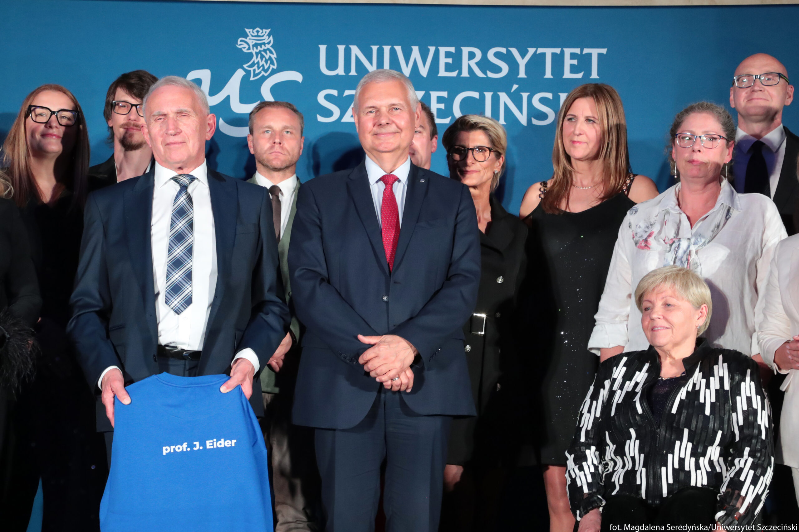 Grupa osób podczas oficjalnego wydarzenia na tle tablicy z napisem „Uniwersytet Szczeciński”. W centrum stoi starszy mężczyzna o siwych włosach, w granatowym garniturze, jasnoniebieskiej koszuli i czerwonym krawacie; ma splecione dłonie przed sobą i lekki uśmiech. Po jego lewej stronie (z perspektywy zdjęcia) stoi inny starszy mężczyzna o siwych włosach, również w granatowym garniturze, z białą koszulą i krawatem w granatowo-szary wzór w kratę; trzyma w dłoniach niebieską koszulkę lub materiał z białym napisem „prof. J. Eider”. Wokół nich stoją inne osoby – kobiety i mężczyźni w średnim i starszym wieku – ubrane elegancko, głównie w ciemne garnitury, sukienki i żakiety; część osób ma okulary. Po prawej stronie na pierwszym planie siedzi starsza kobieta o krótkich, jasnych włosach, w czarno-białej błyszczącej marynarce ze wzorem z prostokątnych elementów; uśmiecha się lekko. W tle widoczny jest niebieski baner z białym logo i napisem „UNIWERSYTET SZCZECIŃSKI”, a pod spodem w prawym dolnym rogu mały biały napis „fot. Magdalena Seredyńska/Uniwersytet Szczeciński”.