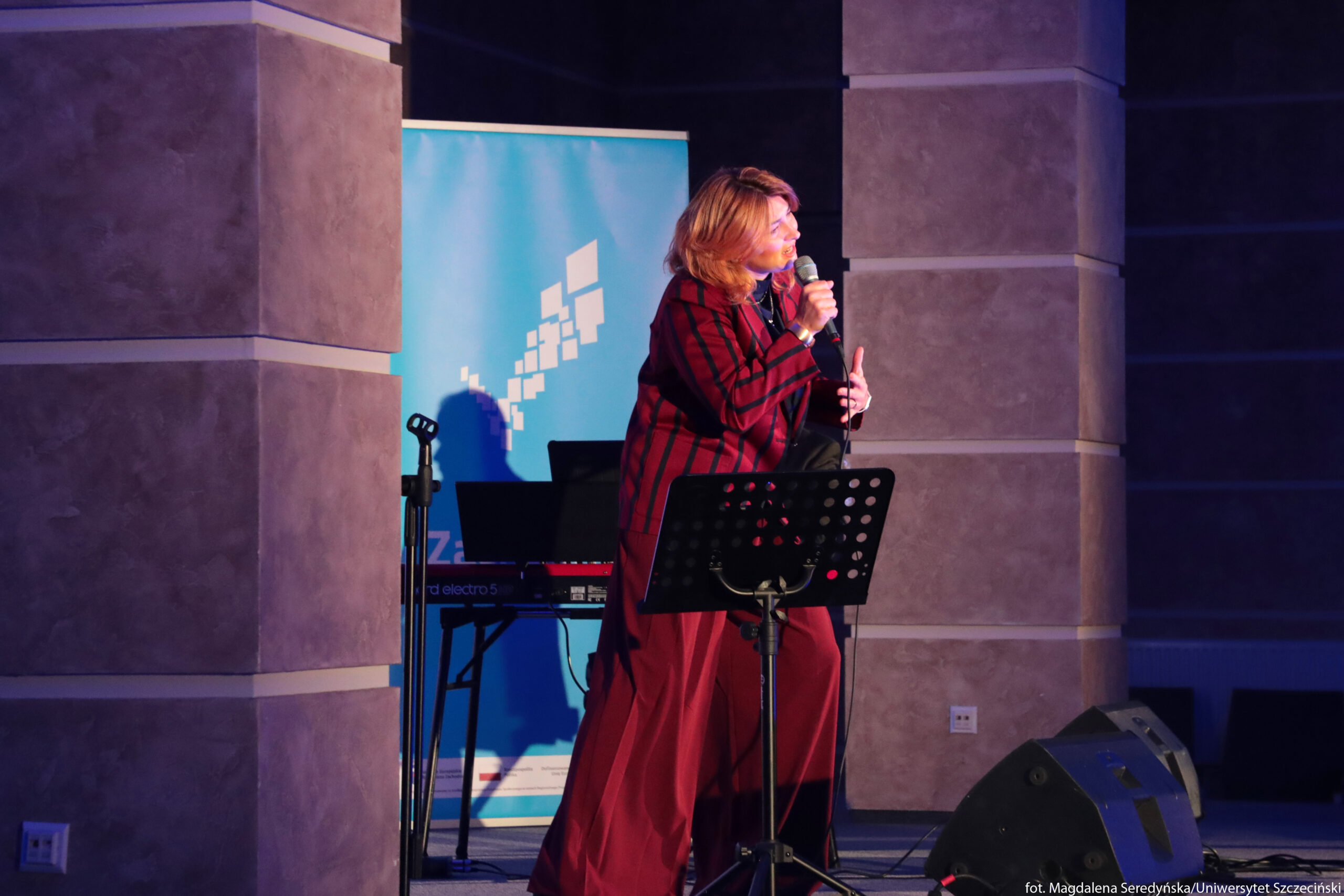 Kobieta występująca na scenie z mikrofonem w dłoni. Stoi między dwoma szerokimi filarami, zwrócona lekko w prawo, w średnim wieku, o jasnobrązowych, sięgających ramion włosach i jasnej skórze. Ma na sobie długi, luźny komplet w odcieniach bordo z pionowymi, ciemniejszymi paskami – marynarkę i bardzo szerokie spodnie sięgające ziemi. Przed nią stoi statyw z czarnym pulpitem z licznymi okrągłymi otworami, służący jako podstawka na nuty lub tekst. W tle widoczna jest jasnoniebieska plansza z białym graficznym motywem złożonym z małych kwadratów, za którą stoi klawiatura elektroniczna na czarnym statywie. Scena jest oświetlona fioletowo-niebieskim światłem, które podkreśla postać i tworzy jej wyraźny cień na niebieskim tle. Na podłodze po prawej stronie leżą dwa czarne głośniki sceniczne skierowane w stronę artystki.