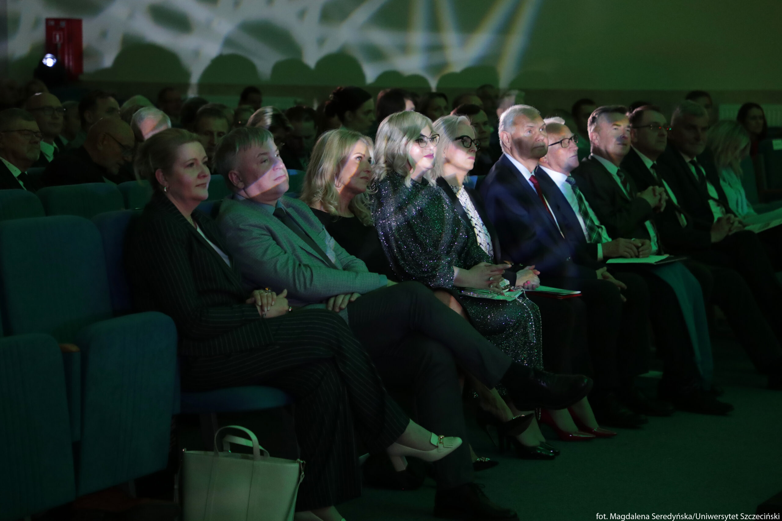 Rząd osób siedzących w sali audytoryjnej. W pierwszym rzędzie siedzi około dziesięciu dorosłych kobiet i mężczyzn w eleganckich, wieczorowych lub biznesowych strojach – garniturach, marynarkach i sukienkach, część w okularach. Wszyscy zwróceni są twarzami w tym samym kierunku, jakby oglądali występ lub prezentację. Na ich twarzach i ubraniach widać jasnozielone i białe refleksy o nieregularnych kształtach, pochodzące z oświetlenia scenicznego. Krzesła mają miękkie, turkusowe obicia. Na podłodze przy pierwszym rzędzie stoi duża, jasna torebka. W tle, w dalszych rzędach, siedzi wiele innych osób, ale są słabiej oświetlone i mniej wyraźne. Cała scena ma przygaszone, teatralne oświetlenie.