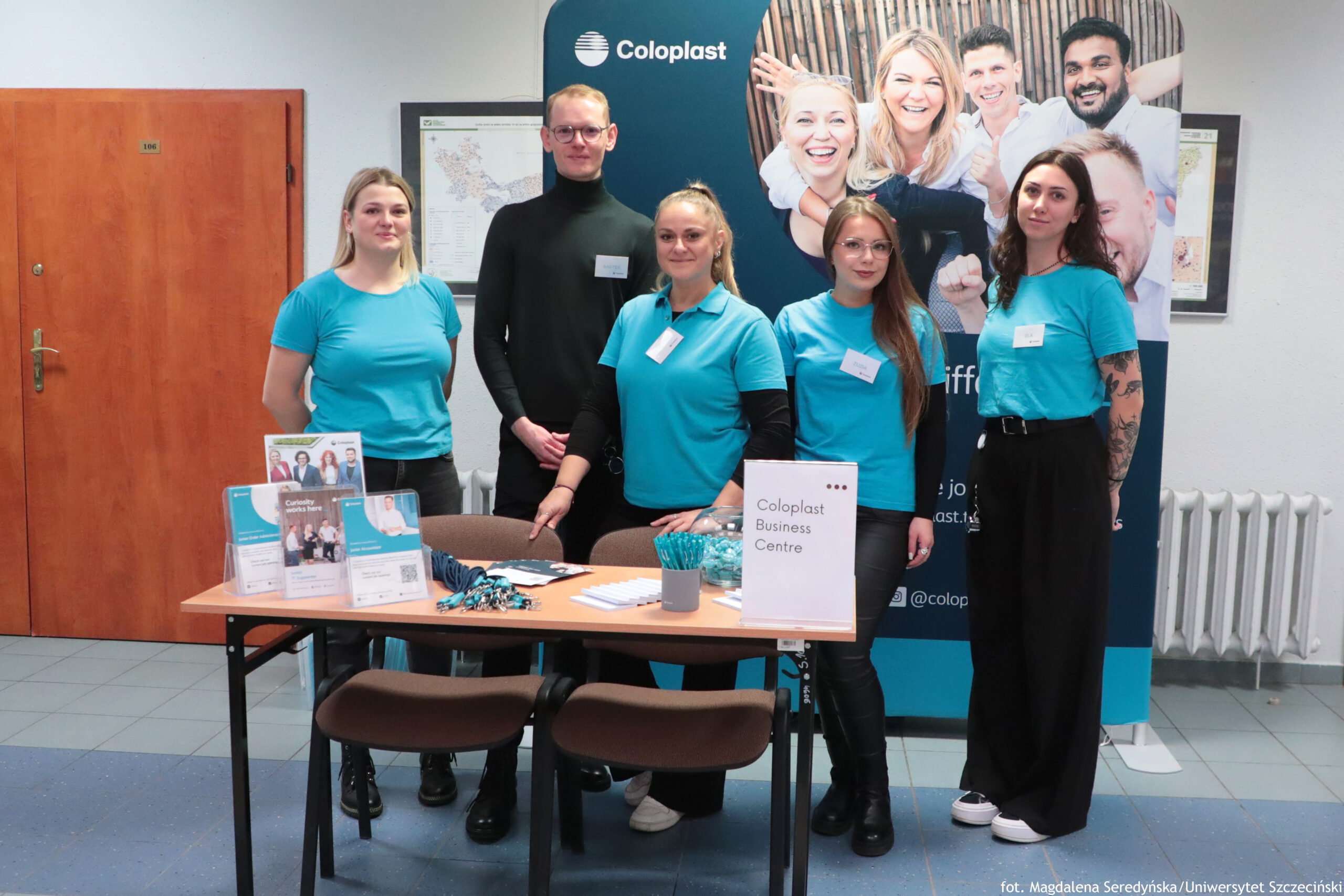 Grupa pięciu osób stojących za stołem promocyjnym z materiałami firmy Coloplast. W tle znajduje się duży stojak reklamowy z napisem „Coloplast” oraz zdjęciami uśmiechniętych ludzi. Cztery kobiety stoją po bokach i z przodu, a mężczyzna stoi pośrodku z tyłu. Większość osób ma na sobie turkusowe koszulki polo z identyfikatorami; mężczyzna ma czarny golf i również identyfikator. Jedna z kobiet po prawej stronie ma widoczne tatuaże na przedramieniu i czarne, szerokie spodnie. Na stole leżą ulotki i stojaki informacyjne, smycze/pendrive’y lub drobne gadżety, długopisy oraz przezroczysta miska z cukierkami. Na jednym z pionowych stojaków na stole widoczny jest napis „Coloplast Business Centre”. W lewym tle widać drewniane drzwi, a po prawej kaloryfer i fragmenty ściennych plansz/map. W prawym dolnym rogu zdjęcia jest podpis: „fot. Magdalena Seredyńska/Uniwersytet Szczeciński”.