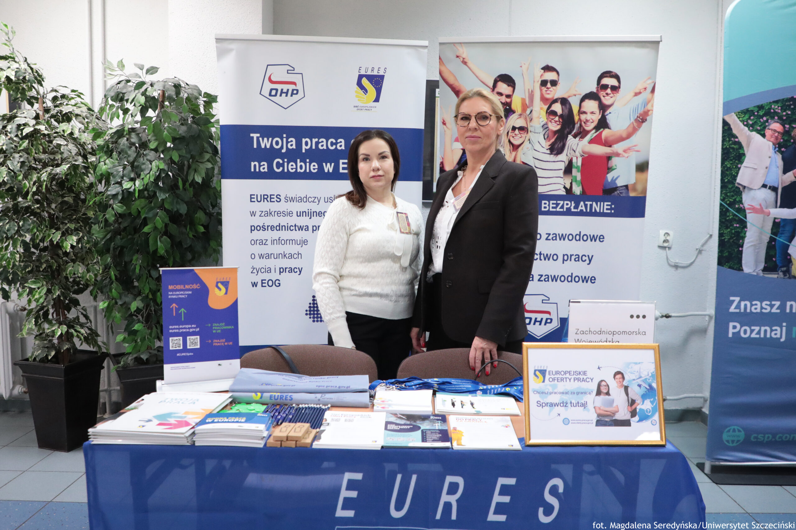 Stoisko informacyjne z dwoma kobietami stojącymi za stołem. Kobieta po lewej ma jasną cerę, ciemne włosy do ramion i białą sweterkową bluzę; kobieta po prawej ma jasną cerę, blond włosy związane z tyłu, okulary oraz czarną marynarkę. Na stole leży dużo materiałów promocyjnych: broszury, ulotki, długopisy, smycze i drobne gadżety; z przodu na niebieskim obrusie widoczny jest duży napis „EURES”. Po prawej na stole stoi ramka z napisem „EUROPEJSKIE OFERTY PRACY” oraz „Sprawdź tutaj!” i zdjęciem dwóch osób.
