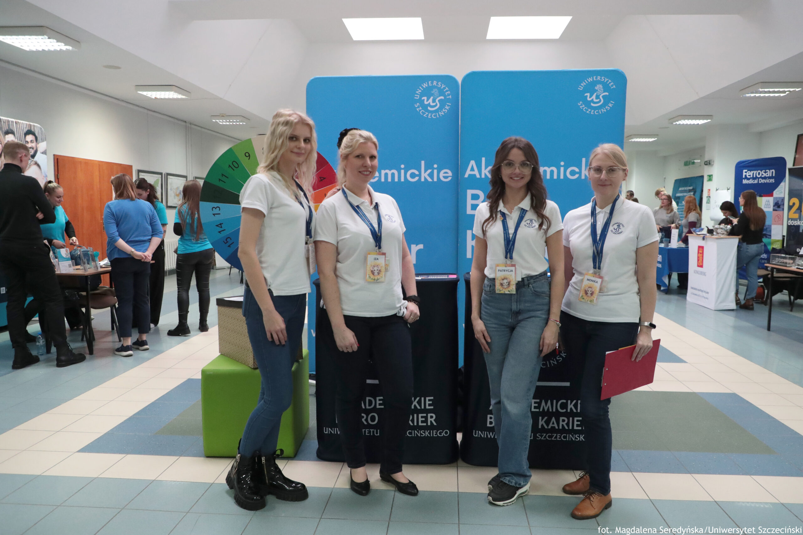 Cztery młode kobiety w białych koszulkach polo stojące na tle niebieskiego stoiska promocyjnego. To zdjęcie wygląda na wydarzenie w budynku (korytarz lub hol) z jasnym oświetleniem sufitowym. Kobiety mają identyfikatory na smyczach; dwie mają długie włosy (jedna blond, jedna brązowe), jedna ma blond włosy spięte, jedna ma jasne blond włosy i okulary. Ubrane są w jeansy lub ciemne spodnie; skrajna z lewej ma czarne, wysokie buty, skrajna z prawej trzyma czerwoną teczkę. Za nimi widać niebieskie ścianki z logo i napisem.