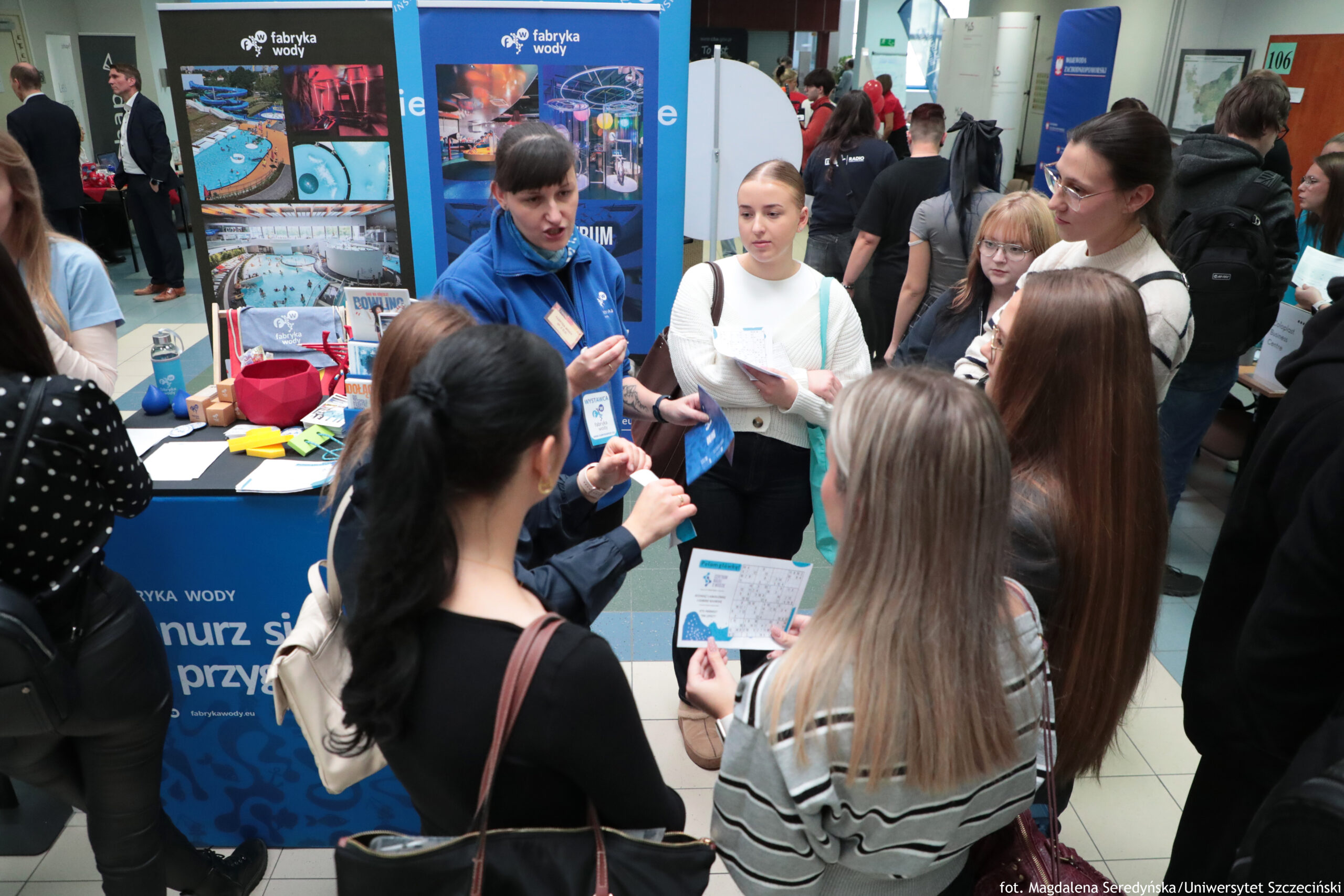 Stoisko informacyjne na targach lub wydarzeniu w budynku, z niebieskimi bannerami „fabryka wody” i grupą młodych osób stojących blisko siebie. W centrum stoi kobieta w niebieskiej bluzie z identyfikatorem na szyi; wygląda na osobę prezentującą ofertę i rozmawiającą z kilkoma uczestniczkami. Wokół niej jest kilka kobiet (młode dorosłe), część trzyma ulotki lub kartki; jedna ma jasny sweter i torebkę na ramieniu, inna ma okulary i jasne włosy z grzywką. Na pierwszym planie widać tyły głów kilku osób oraz torby na ramieniu. Za ladą stoiska leżą różne materiały: ulotki, drobne gadżety i pojemniki, a na tle stoją duże plansze ze zdjęciami obiektów wodnych i instalacji (baseny, zjeżdżalnie, elementy parku wodnego). W tle po bokach widać więcej stoisk i ludzi w korytarzu.