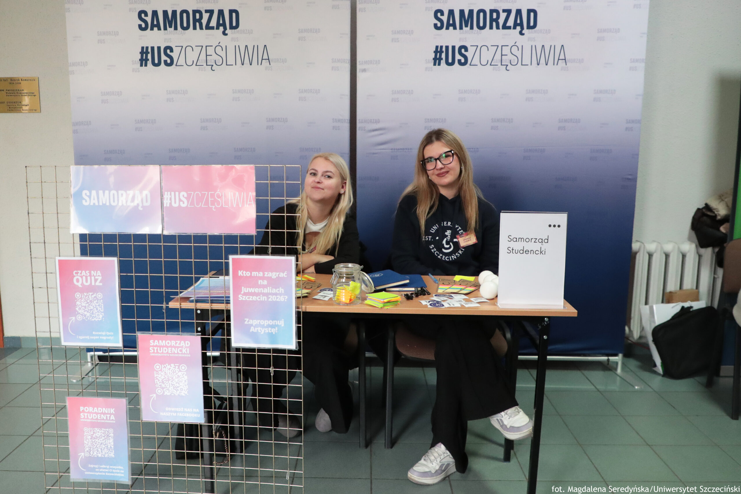 Stanowisko informacyjne Samorządu Studenckiego z dwoma siedzącymi kobietami za stołem. W tle są dwie duże ścianki z napisem „SAMORZĄD” oraz hasztagiem „#USZCZĘŚLIWIA”. Na kratownicy obok stołu wiszą kolorowe plakaty, część z kodami QR. Osoby za stołem: Kobieta po lewej: jasne, długie blond włosy, jasna skóra, ciemna bluza; siedzi lekko bokiem i patrzy w stronę aparatu. Kobieta po prawej: jasne, długie włosy (ciemny blond/jasny brąz), jasna skóra, czarne okulary, ciemna bluza z nadrukiem; uśmiecha się.