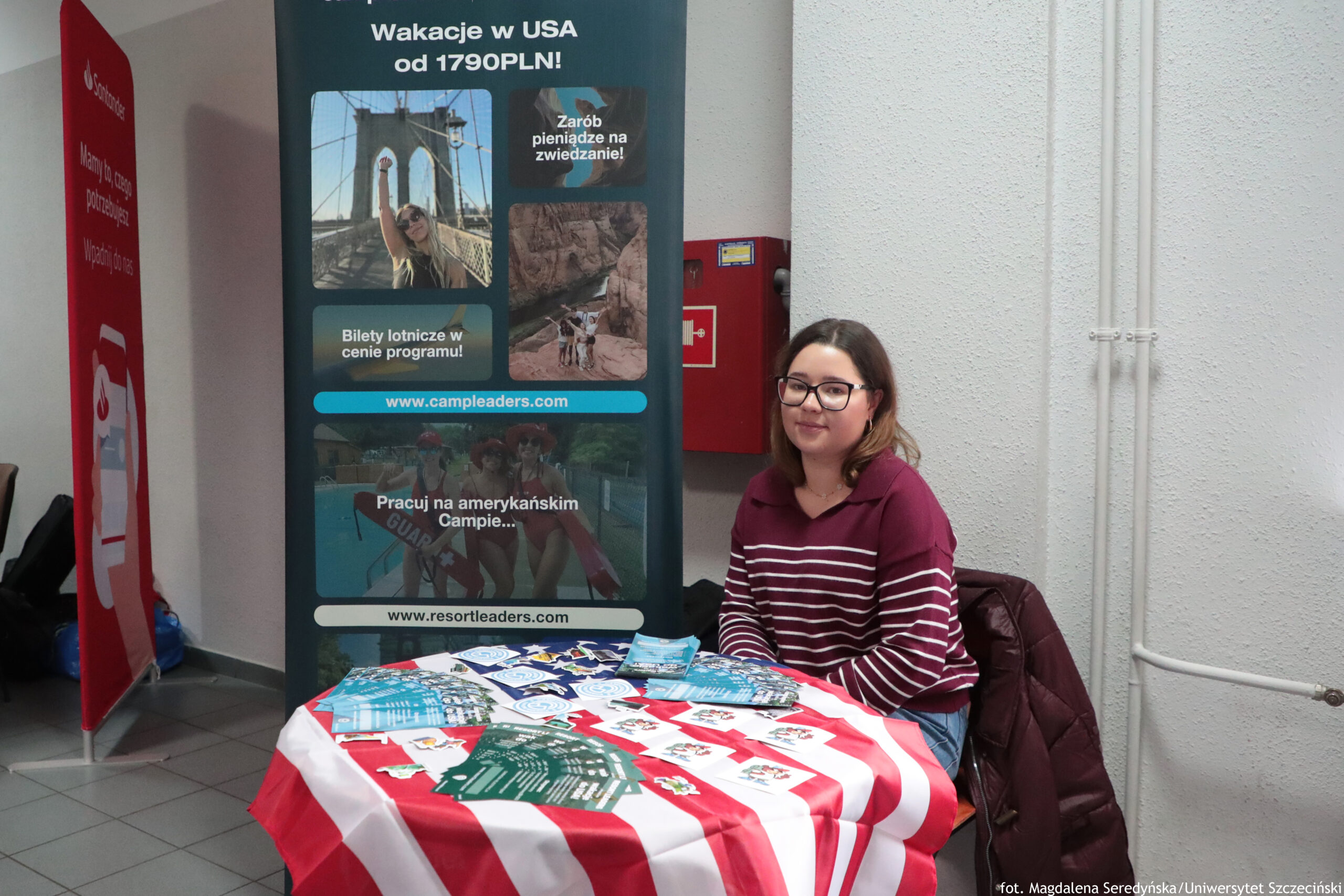 Stoisko informacyjne z siedzącą kobietą przy stole przykrytym czerwono-białym obrusem w pasy. Na stole leżą rozłożone ulotki i broszury.Kobieta ma jasną cerę, ciemnobrązowe włosy do ramion i czarne okulary. Ma na sobie bordowy sweter w białe poziome paski i siedzi przy stoliku; obok leży bordowa kurtka na krześle. W tle widać jasną ścianę z rurkami instalacyjnymi oraz czerwone skrzynki (prawdopodobnie przeciwpożarowe/medyczne). Po lewej stronie kadru stoi czerwony pionowy baner. W prawym dolnym rogu jest podpis: „fot. Magdalena Seredyńska/Uniwersytet Szczeciński”.