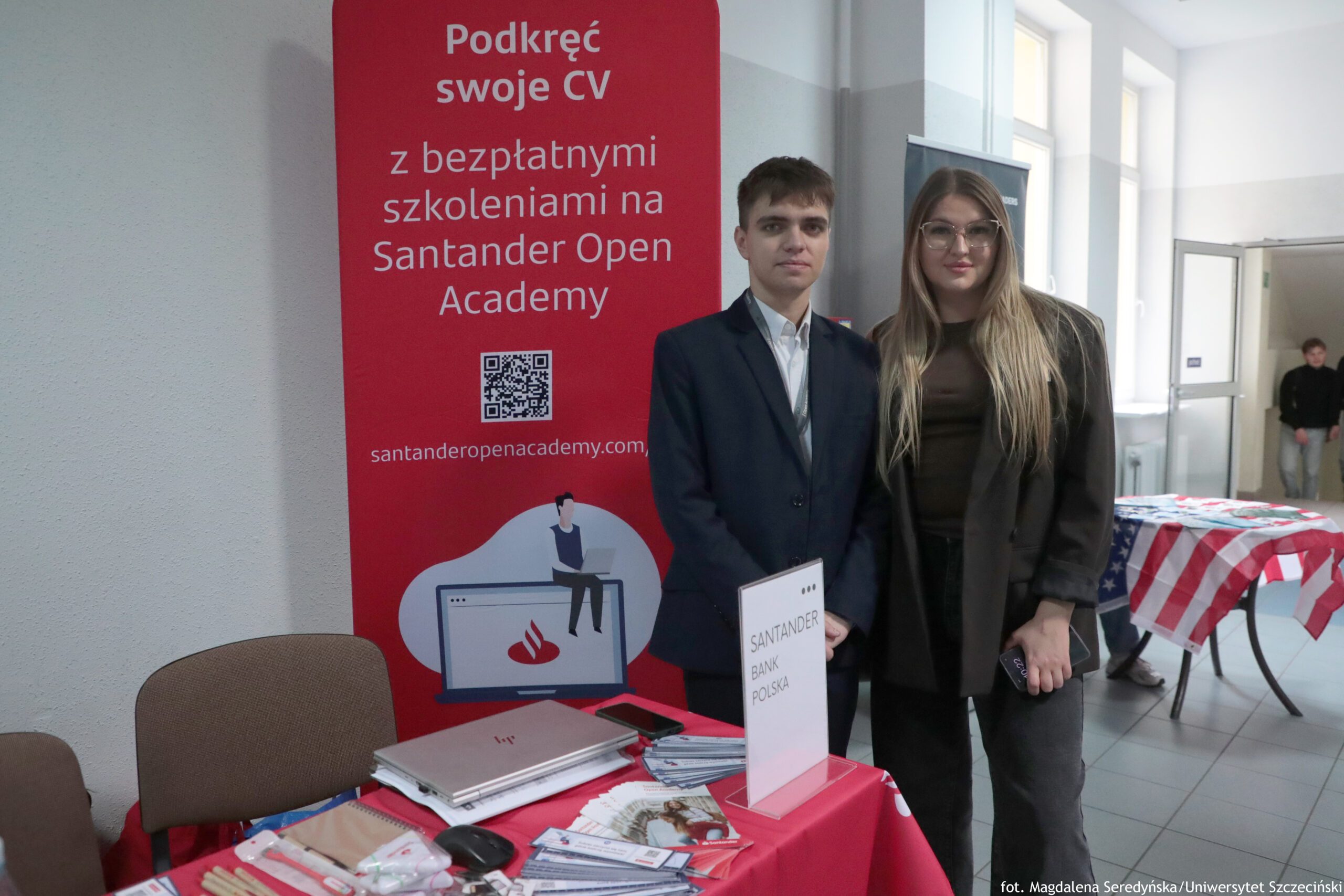 Stoisko promocyjne Santander z czerwonym banerem i dwiema osobami stojącymi przed nim w korytarzu budynku. Na banerze widoczny jest napis: „Podkręć swoje CV z bezpłatnymi szkoleniami na Santander Open Academy”. Na dole jest grafika laptopa z logo Santander i postacią siedzącą na ekranie. Dwie młode osoby stoją obok siebie: mężczyzna w ciemnej marynarce i jasnej koszuli oraz kobieta w okularach, z długimi jasnymi włosami, w ciemnym ubraniu i luźnej marynarce. Na stole przed nimi leżą materiały promocyjne (ulotki, długopisy i gadżety), laptop, mysz oraz stojak z kartką „SANTANDER BANK POLSKA”. W tle po prawej widać drzwi i fragment innego stołu z czerwono-biało-niebieskim obrusem.