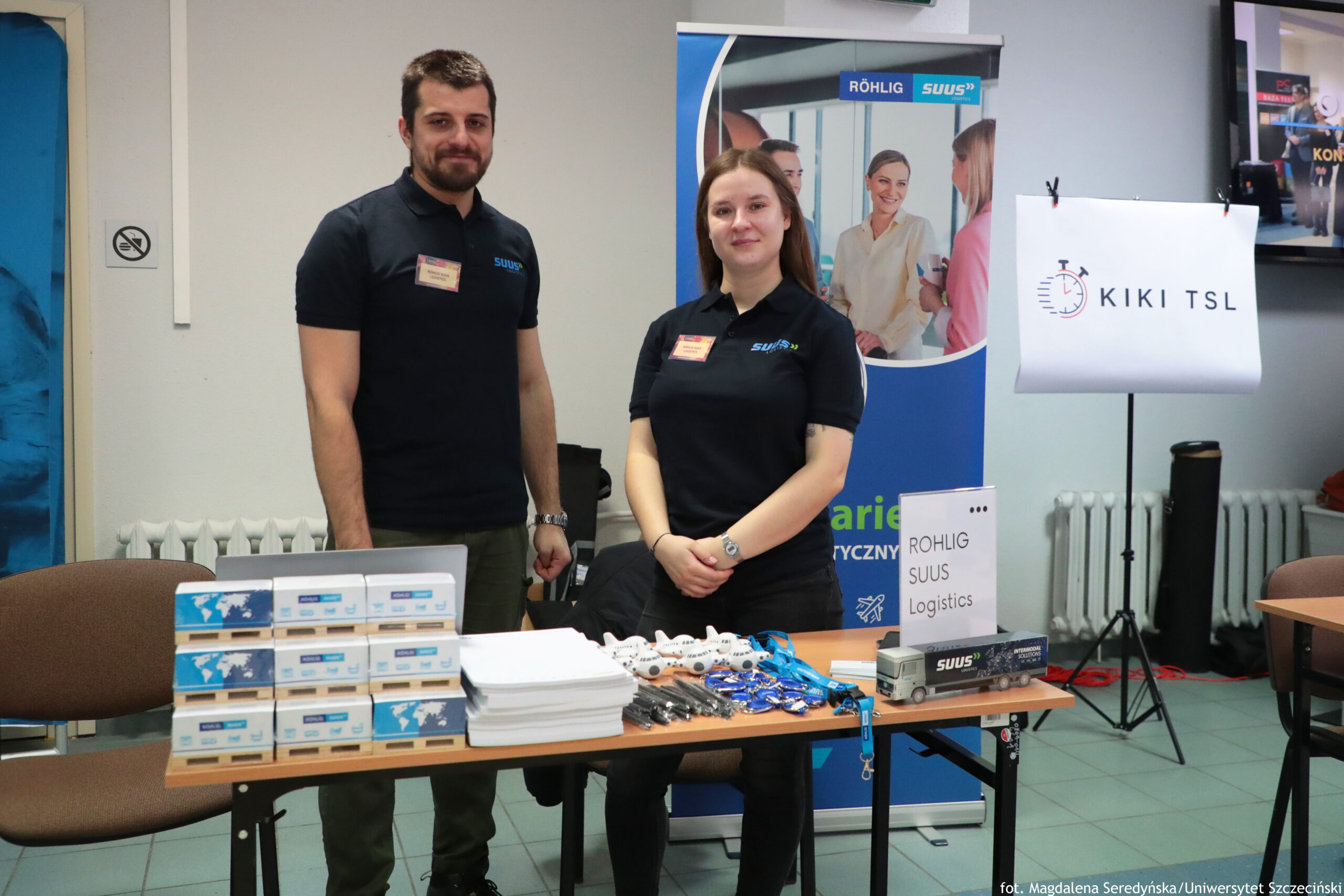 Stoisko promocyjne w holu, za którym stoją dwie osoby w czarnych koszulkach polo z logo firmy. Na stole leżą ułożone w stosy pudełka, plik białych kartek lub notesów, długopisy oraz niebieskie smycze i drobne gadżety. W tle widać roll-up z napisem „RÖHLIG SUUS” oraz fotografią ludzi w biurze, a po prawej kartkę na stojaku z napisem „KIKI TSL”. Na małej tabliczce na stole jest tekst „ROHLIG SUUS Logistics”.