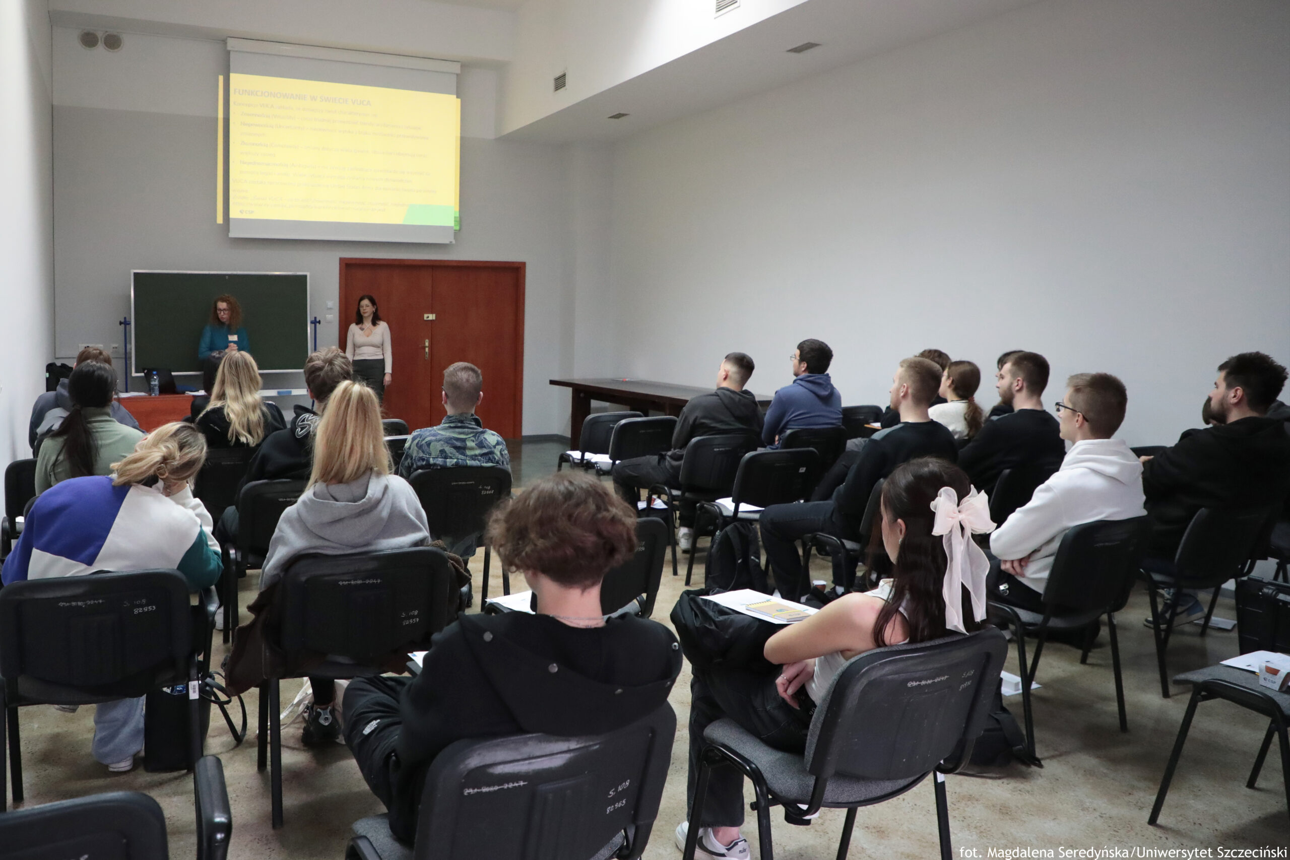Sala wykładowa z publicznością siedzącą na czarnych krzesłach, skierowaną w stronę przodu sali. Na przodzie są dwie osoby prowadzące: jedna stoi przy stole i tablicy, druga stoi obok czerwonych drzwi. Nad nimi wisi ekran z wyświetloną prezentacją. Widzowie to głównie młodzi dorośli; część ma notatki lub kartki na kolanach. Na dole po prawej stronie widać podpis fotograficzny: „fot. Magdalena Seredyńska/Uniwersytet Szczeciński”.