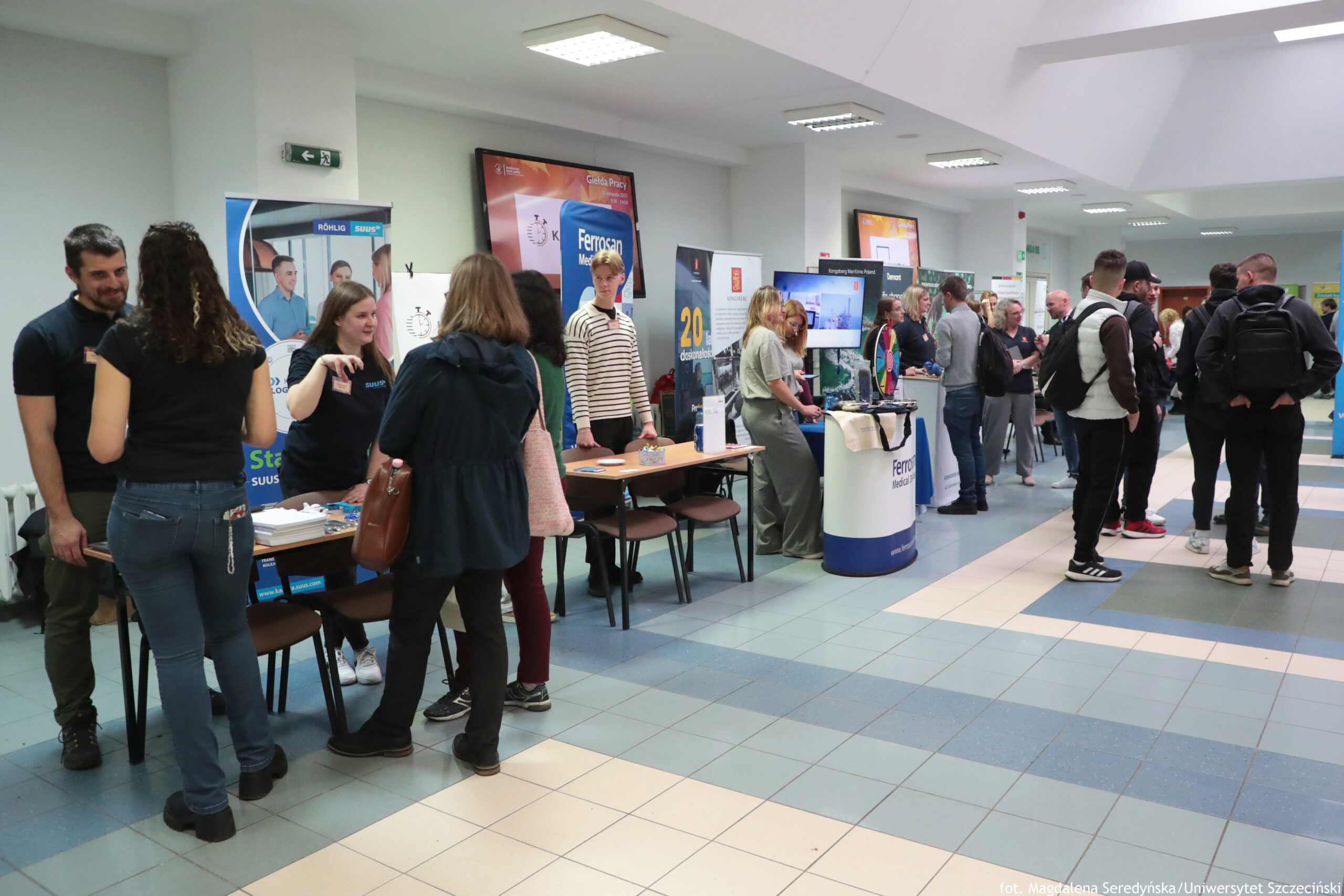 Hol z targami, z kilkoma stoiskami informacyjnymi ustawionymi w jednym rzędzie. W pomieszczeniu jest jasne oświetlenie sufitowe i podłoga w niebiesko-białe płytki. Po lewej stronie widać stół z materiałami promocyjnymi; stoją przy nim osoby rozmawiające, głównie młodzi dorośli w codziennych ubraniach (jeansy, bluzy, kurtki). Za stołami stoją pionowe roll-upy z grafikami i logotypami firm/instytucji. Na środku i w głębi jest więcej stoisk z banerami; część osób stoi w małych grupach i rozmawia z przedstawicielami stoisk. Na ścianie u góry po lewej jest zielony znak wyjścia ewakuacyjnego. W prawym dolnym rogu zdjęcia znajduje się mały podpis: „fot. Magdalena Seredyńska/Uniwersytet Szczeciński”.