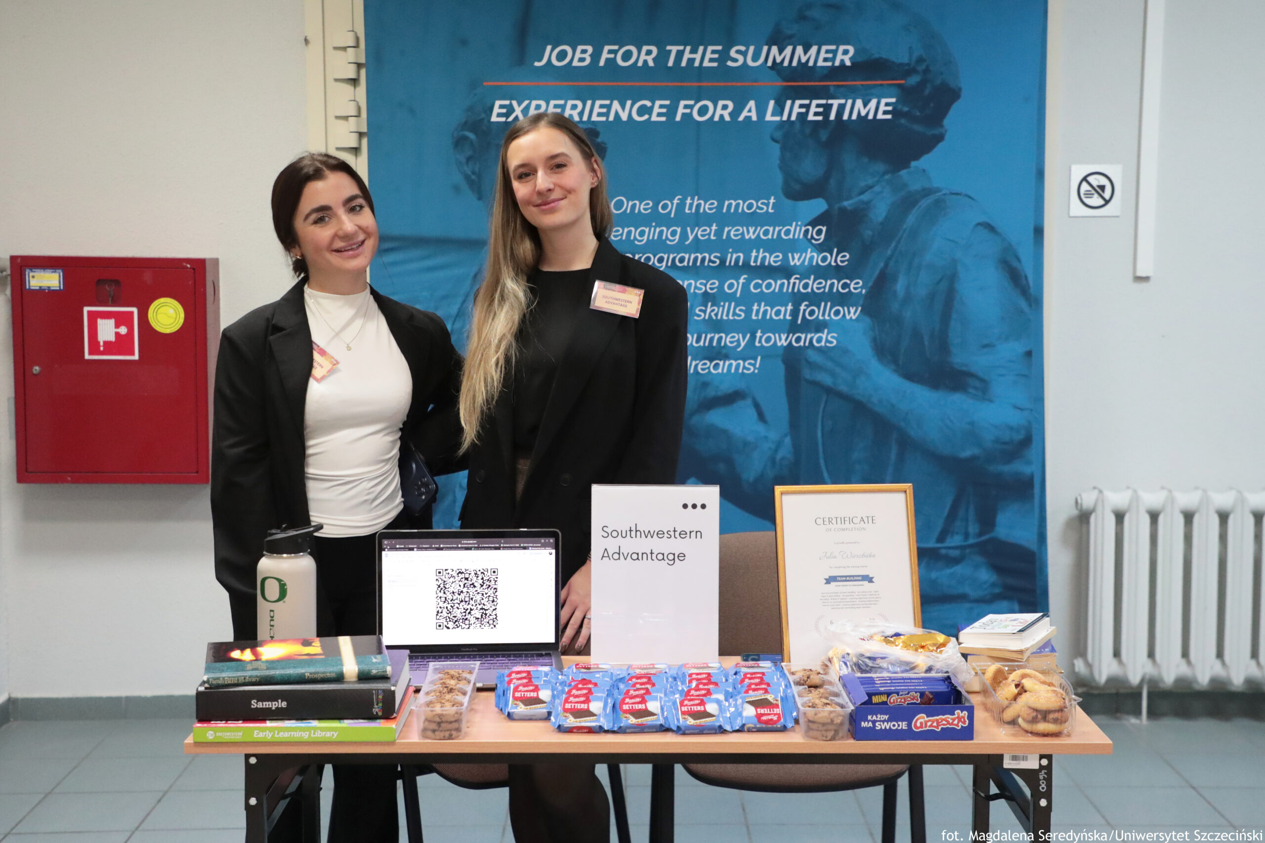 Dwa młode kobiety stoją za stołem informacyjnym na korytarzu. Na stole leżą przekąski i materiały promocyjne: otwarty laptop z dużym kodem QR na ekranie, stojąca kartka z napisem „Southwestern Advantage”, oprawiony dokument z nagłówkiem „CERTIFICATE OF COMPLETION”, kilka książek, butelka/bidon z logo „O”, plastikowe pojemniki z ciastkami oraz rząd niebieskich paczek słodyczy. Kobieta po lewej ma ciemne włosy związane z tyłu, jasną cerę, biały dopasowany top i czarną marynarkę. Kobieta po prawej ma długie jasne blond włosy, jasną cerę i czarną marynarkę; obie mają przypięte identyfikatory. W tle wisi duży niebieski plakat z czytelnym tekstem: „JOB FOR THE SUMMER” oraz „EXPERIENCE FOR A LIFETIME”. Po lewej na ścianie jest czerwona skrzynka przeciwpożarowa, a po prawej znak zakazu palenia. Na dole po prawej widoczny napis: „fot. Magdalena Seredyńska/Uniwersytet Szczeciński”.