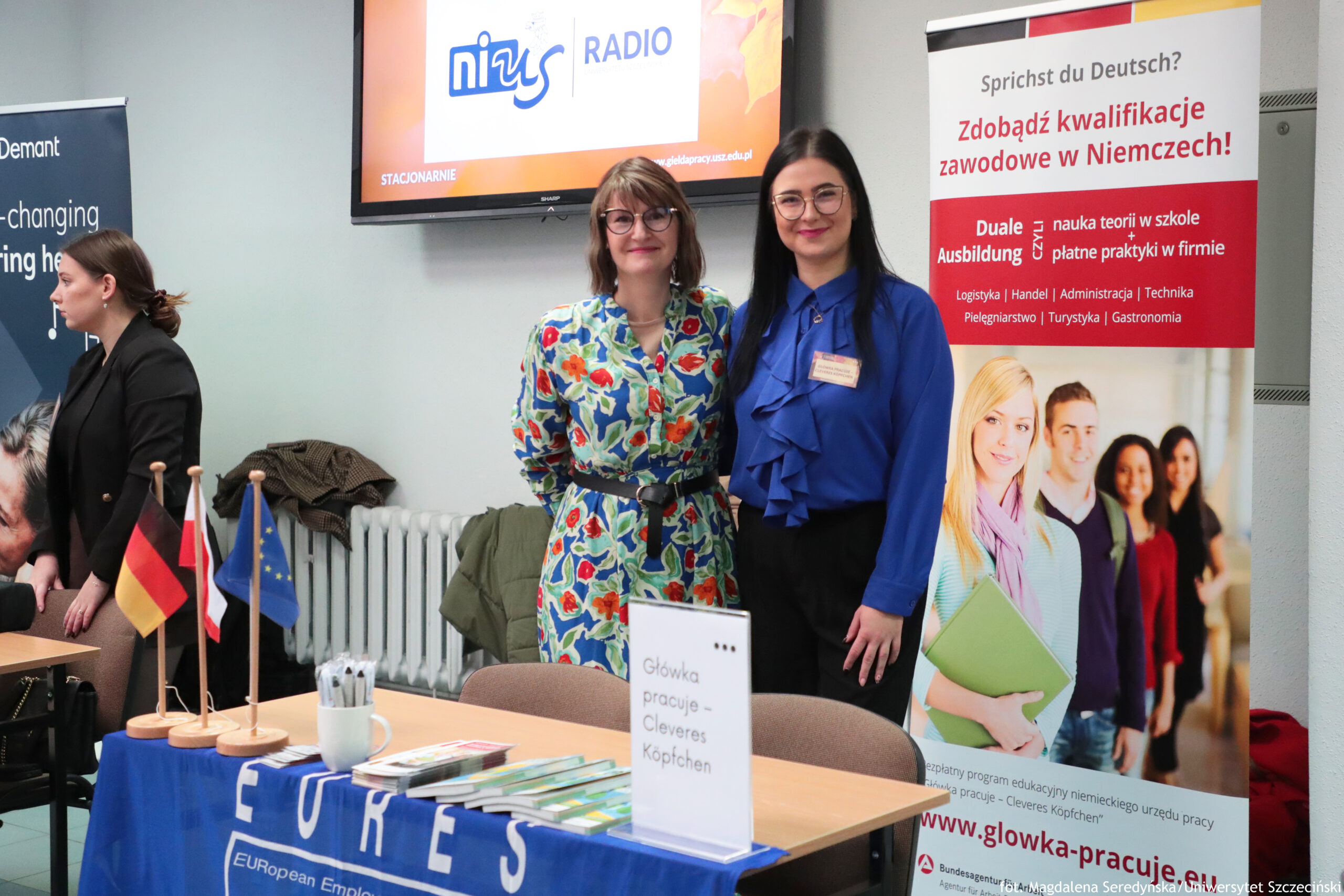 Stoisko informacyjne w sali, z dwoma kobietami stojącymi obok siebie i pozującymi do zdjęcia. Kobieta po lewej ma krótkie, jasnobrązowe włosy, okulary i kolorową sukienkę we wzory (niebieskie tło z czerwono-zielonymi motywami) z czarnym paskiem. Kobieta po prawej ma długie, ciemne włosy, okulary i granatową/bluową bluzkę z falbaną z przodu oraz czarne spodnie; na piersi ma identyfikator.