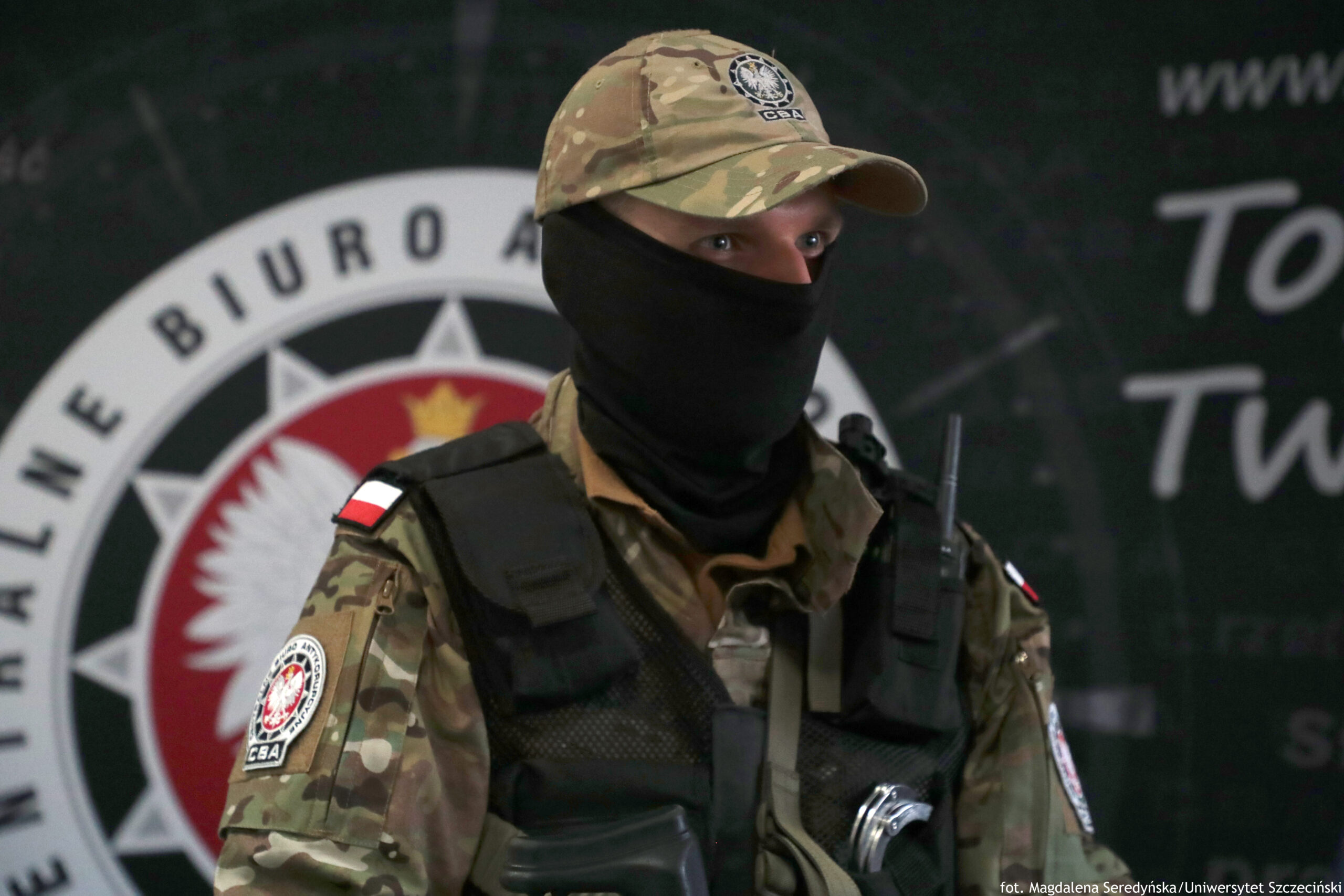 Zamaskowana osoba w mundurze moro i czarnej kamizelce taktycznej, w czapce z daszkiem i czarnej kominiarce zasłaniającej dolną część twarzy. Widoczne są jasne oczy oraz fragment jasnej skóry wokół nich. Na ramionach znajdują się naszywki z polską flagą. Na lewym rękawie jest okrągła naszywka z godłem i napisem „CBA”. Na czapce także widać emblemat „CBA”.
