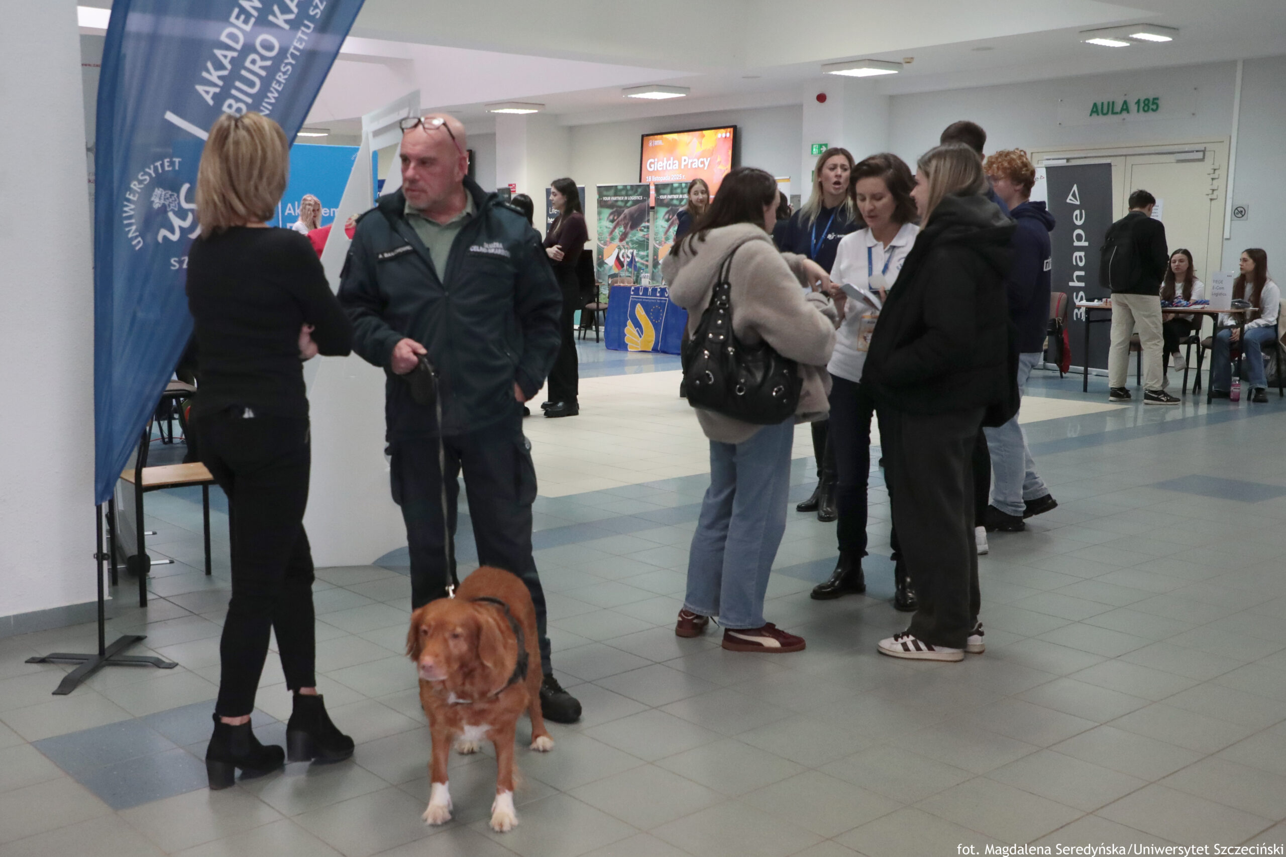 Hol budynku uczelni z kilkunastoma osobami podczas wydarzenia informacyjnego. Na pierwszym planie stoi mężczyzna w ciemnej kurtce i spodniach, obok niego rudy pies na smyczy. Po lewej stronie widać wysoką niebieską flagę reklamową. W tle stoją grupki ludzi rozmawiających przy stoiskach; na jednym ekranie wyświetla się napis „Giełda Pracy”. Po prawej na ścianie jest zielony napis „AULA 185”, a obok stoi pionowy baner z napisem „3shape”. Na dole zdjęcia widnieje podpis: „fot. Magdalena Seredyńska/Uniwersytet Szczeciński”.