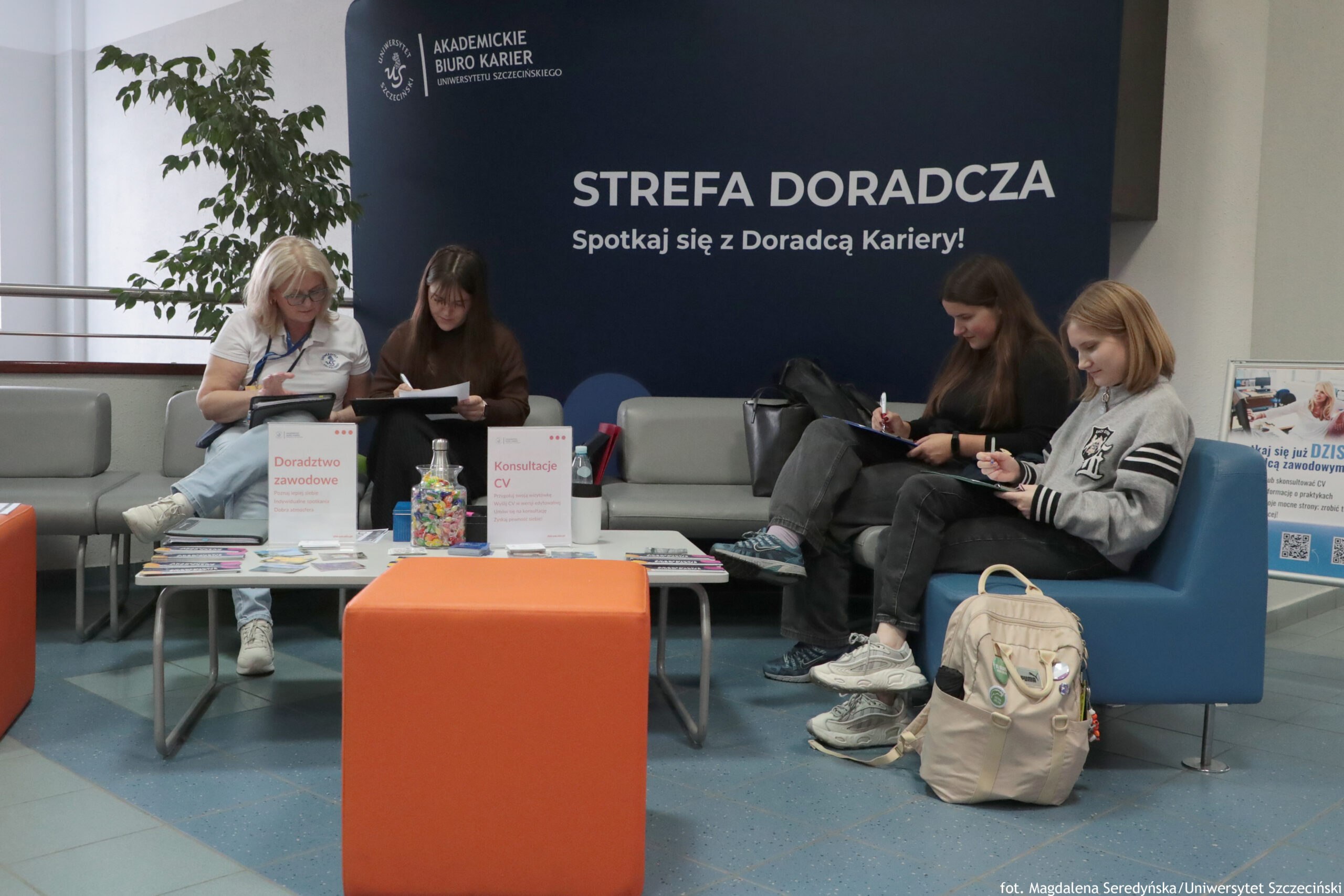 Strefa doradcza w holu uczelni, z czterema kobietami siedzącymi na kanapach i wypełniającymi formularze na podkładkach. W tle jest duży granatowy baner z białymi napisami: „AKADEMICKIE BIURO KARIER UNIWERSYTETU SZCZECIŃSKIEGO”, duży tytuł „STREFA DORADCZA” oraz „Spotkaj się z Doradcą Kariery!”. Po lewej siedzi starsza kobieta z jasnymi włosami i okularami, w białej koszulce, coś zapisuje. Obok niej siedzi młoda kobieta z długimi ciemnymi włosami, również pisze na kartce. Po prawej stronie na kanapie siedzą dwie młode kobiety: jedna z długimi brązowymi włosami, druga z jasnymi, krótszymi włosami w szarej bluzie; obie notują. Na środku stoi niski stolik z ulotkami i dwoma stojącymi tabliczkami: „Doradztwo zawodowe” oraz „Konsultacje CV”. Na stoliku jest też przezroczysty słoik z kolorowymi cukierkami i kilka drobnych przedmiotów (np. długopisy). Z przodu znajduje się pomarańczowy puf. Na podłodze z prawej strony leży jasny plecak. Po lewej w tle stoi duża roślina doniczkowa. W prawym dolnym rogu widoczny podpis: „fot. Magdalena Seredyńska/Uniwersytet Szczeciński”.