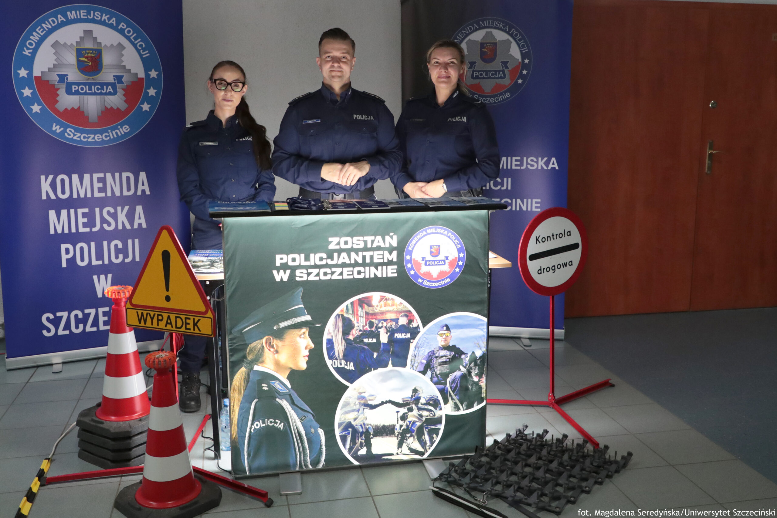Stanowisko informacyjne policji z trzema funkcjonariuszami w granatowych mundurach stojącymi za stołem, na tle bannerów „Komenda Miejska Policji w Szczecinie”. Na środku stoi mężczyzna, po bokach dwie kobiety; wszyscy patrzą w stronę aparatu. Na stole leżą materiały informacyjne. Z przodu stołu jest duży baner z napisem „Zostań policjantem w Szczecinie” oraz zdjęciami policjantów w służbie. Po lewej stronie stoją dwa pachołki drogowe i trójkątny znak ostrzegawczy z napisem „Wypadek”. Po prawej stronie jest okrągły znak „Kontrola drogowa” na czerwonym stojaku. Na podłodze po prawej leży czarny element przypominający matę lub kolczatkę. W tle po prawej widać brązowe drzwi.