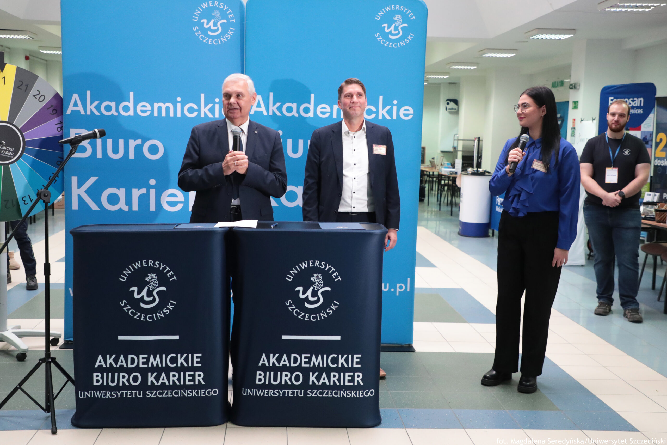 Scena wydarzenia na uczelni z czterema osobami stojącymi przed niebieskimi ściankami z logo „Uniwersytet Szczeciński” i dużym napisem „Akademickie Biuro Karier”. Na pierwszym planie stoją dwa połączone stoliki okryte granatowymi pokrowcami z białym logo i napisem: „UNIWERSYTET SZCZECIŃSKI” oraz „AKADEMICKIE BIURO KARIER UNIWERSYTETU SZCZECIŃSKIEGO”. Po lewej stronie stoi starszy mężczyzna o jasnej skórze, siwych krótkich włosach, w ciemnym garniturze i białej koszuli; trzyma mikrofon w obu dłoniach, jakby przemawiał. Obok niego stoi młodszy mężczyzna o jasnej skórze, krótkich brązowych włosach, w ciemnej marynarce i białej koszuli, z identyfikatorem na piersi. Po prawej stoi młoda kobieta o jasnej skórze, długich ciemnych włosach i okularach, w niebieskiej bluzce i czarnych spodniach; trzyma mikrofon i jest zwrócona w stronę mężczyzn. Najbardziej z prawej stoi mężczyzna o jasnej skórze z brodą, w czarnym T-shircie i z identyfikatorem, z rękami splecionymi przed sobą. W tle widać wnętrze budynku z jasnymi kafelkami na podłodze, oświetleniem sufitowym, stolikami oraz stoiskami/banerami.