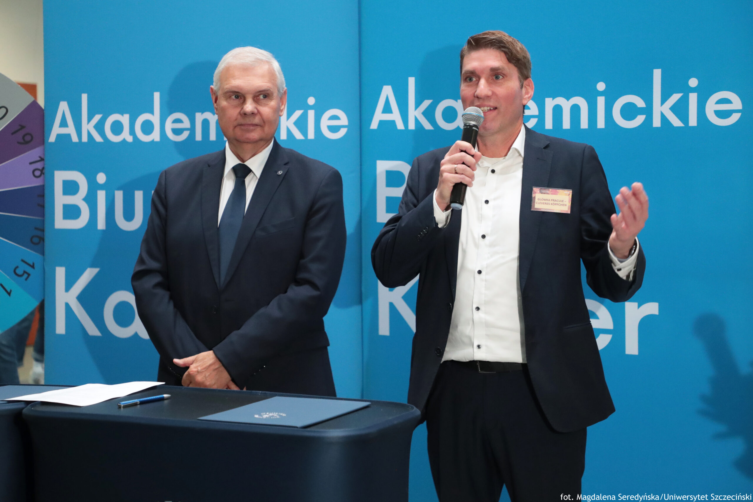 Dwaj mężczyźni w garniturach stoją na tle niebieskiej ścianki z dużymi białymi napisami. Mężczyzna po lewej ma krótkie białe włosy, jasną skórę i ciemny garnitur z białą koszulą oraz ciemnym krawatem. Ma ręce złożone z przodu i patrzy w stronę mężczyzny po prawej. Mężczyzna po prawej ma krótkie brązowe włosy, jasną skórę, ciemny garnitur i białą koszulę bez krawata. Trzyma mikrofon przy ustach i gestykuluje drugą dłonią; na marynarce ma przypięty identyfikator.