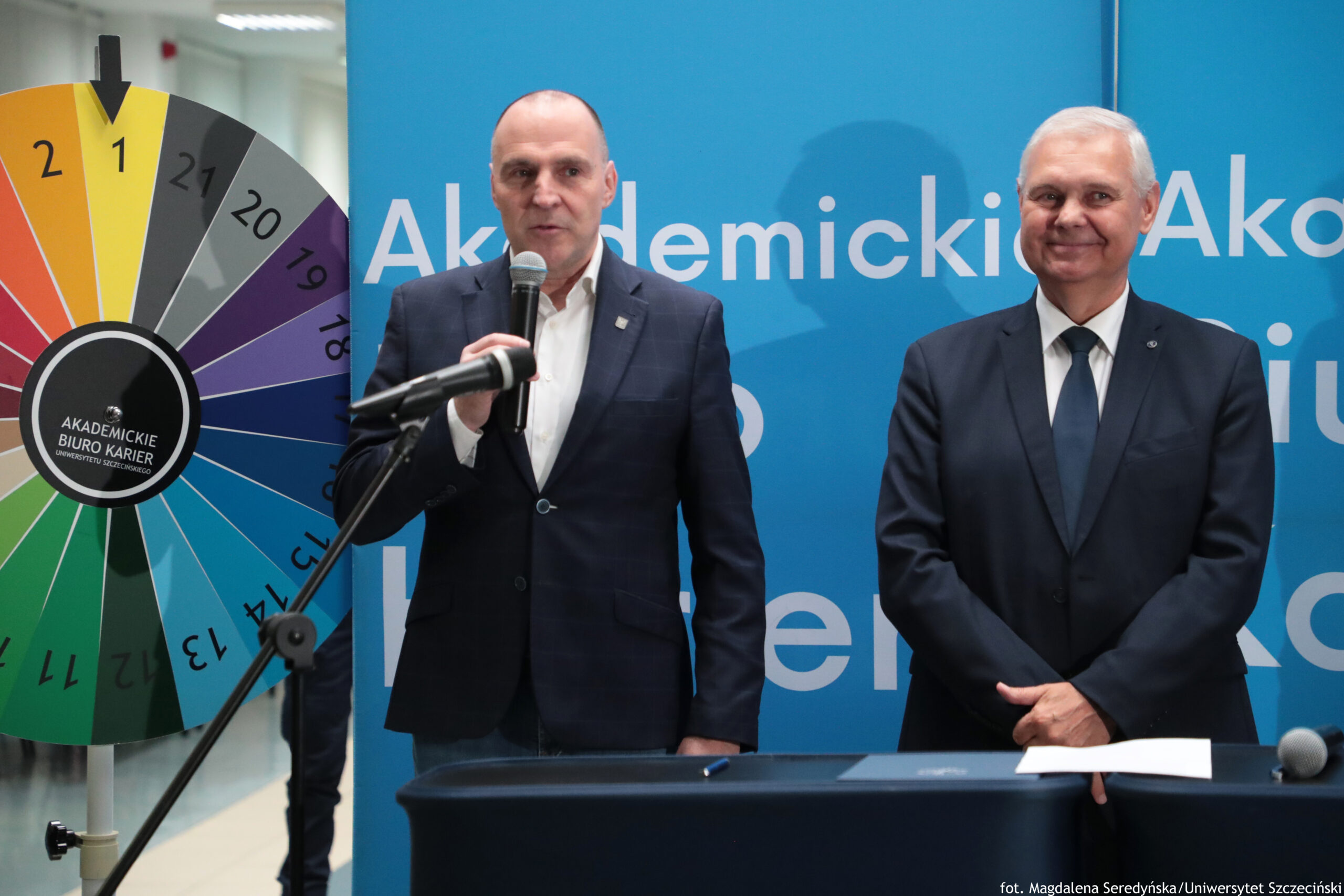 Dwóch mężczyzn w garniturach stojących przy stole podczas wydarzenia lub konferencji. W tle jest duży niebieski baner z białym napisem. Mężczyzna po lewej ma jasną skórę i jest łysy. Trzyma mikrofon i wygląda na osobę przemawiającą. Przed nim widać statyw z mikrofonem skierowanym w jego stronę. Mężczyzna po prawej ma jasną skórę i siwe, krótko ścięte włosy. Stoi z rękami złożonymi, w białej koszuli i ciemnym krawacie, z lekkim uśmiechem. Po lewej stronie kadru znajduje się duże kolorowe koło losujące. Na środku koła jest czarne pole z białym napisem: „AKADEMICKIE BIURO KARIER” oraz mniejszym tekstem „UNIWERSYTETU SZCZECIŃSKIEGO”. Na stole leży kartka papieru i długopis. W prawym dolnym rogu zdjęcia widoczny jest podpis: „fot. Magdalena Seredyńska/Uniwersytet Szczeciński”.