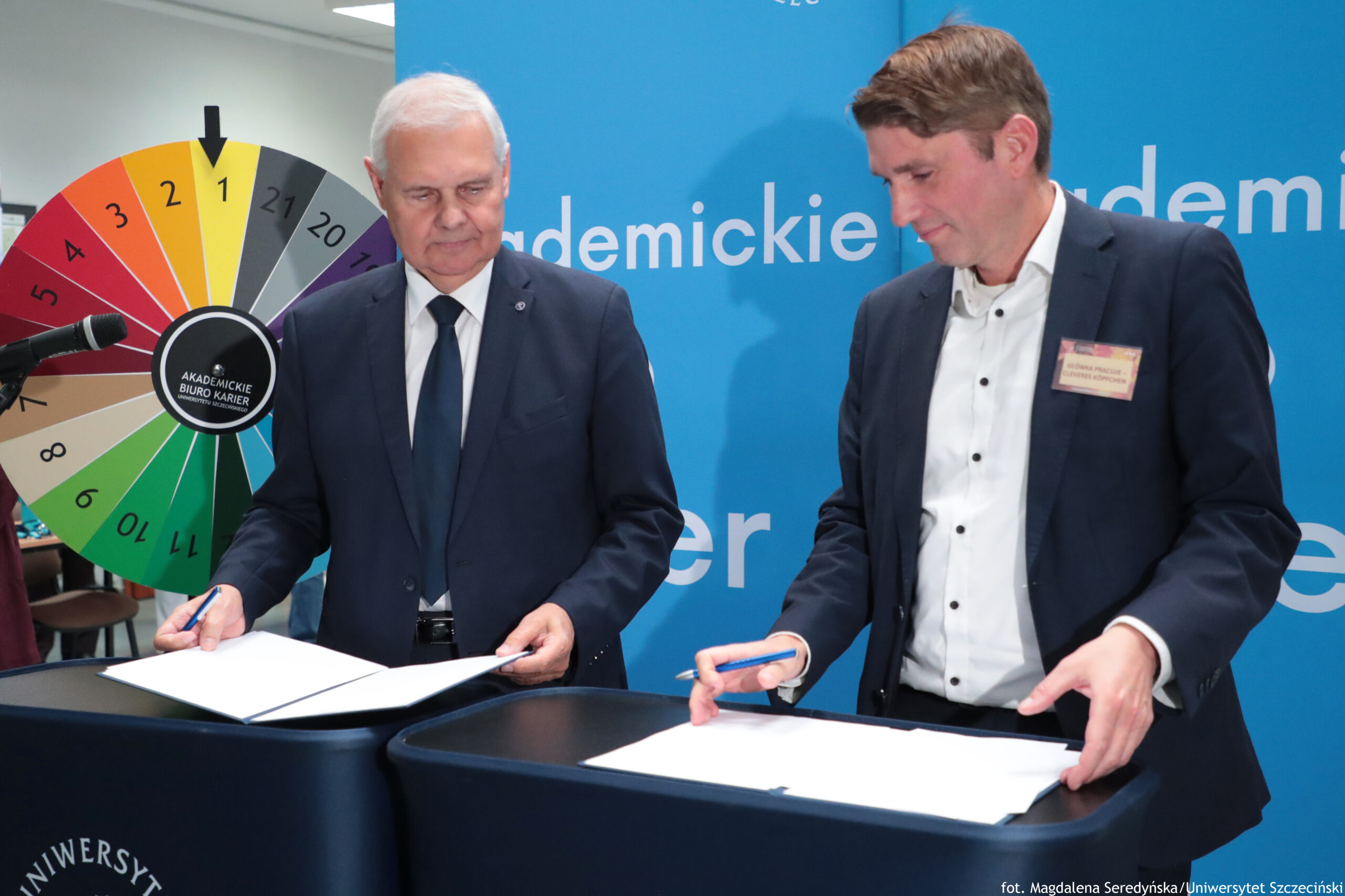 Dwóch mężczyzn w garniturach podpisujących dokumenty na stojących pulpitach. Mężczyzna po lewej ma siwe włosy, białą koszulę i ciemny krawat; mężczyzna po prawej ma brązowe włosy, białą koszulę bez krawata i identyfikator przypięty do marynarki. W tle po lewej stoi kolorowe koło z ponumerowanymi polami (jak koło fortuny) z czarną strzałką wskazującą pole „1”; na środku koła widoczny napis „AKADEMICKIE BIURO KARIER”. Za nimi jest niebieska ścianka z dużym. Po lewej stronie kadru widać mikrofon na statywie. W prawym dolnym rogu jest drobny podpis: „fot. Magdalena Seredyńska/Uniwersytet Szczeciński”.